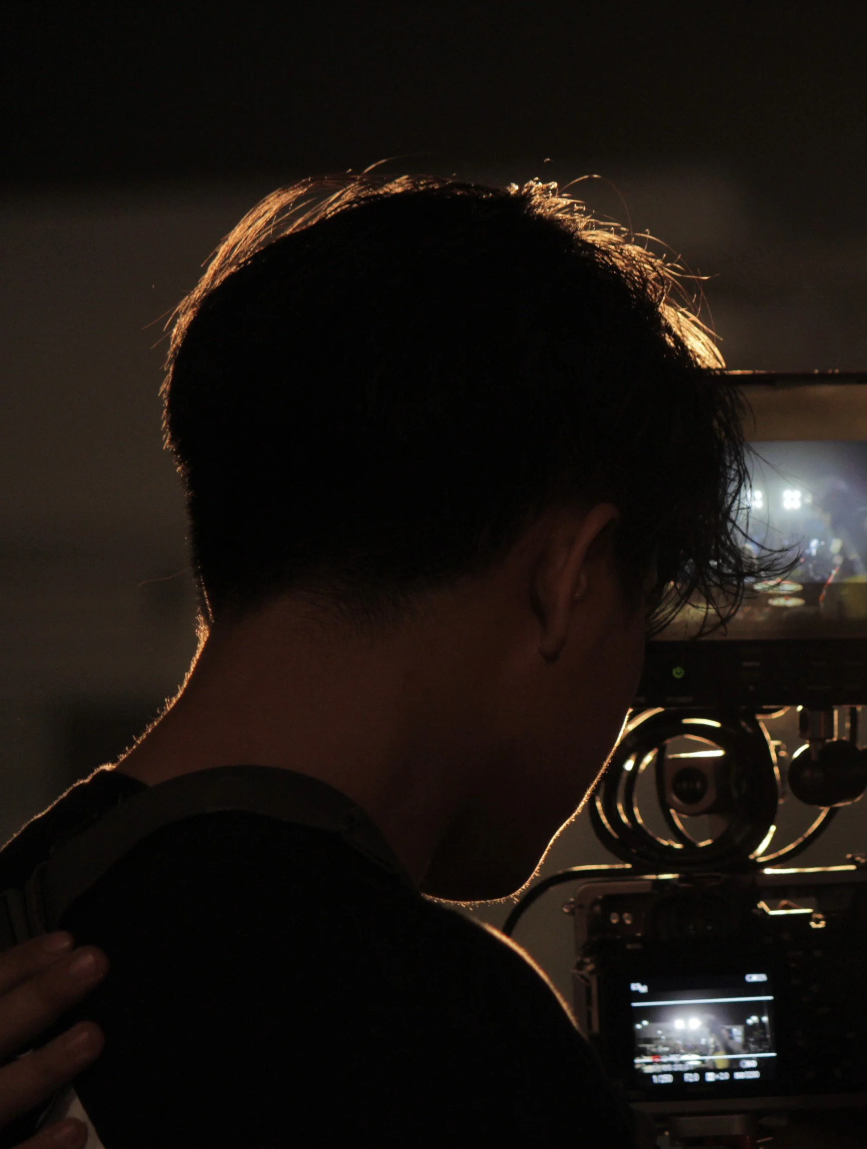 A person with dark hair is filming with a camera, backlit by light from behind.