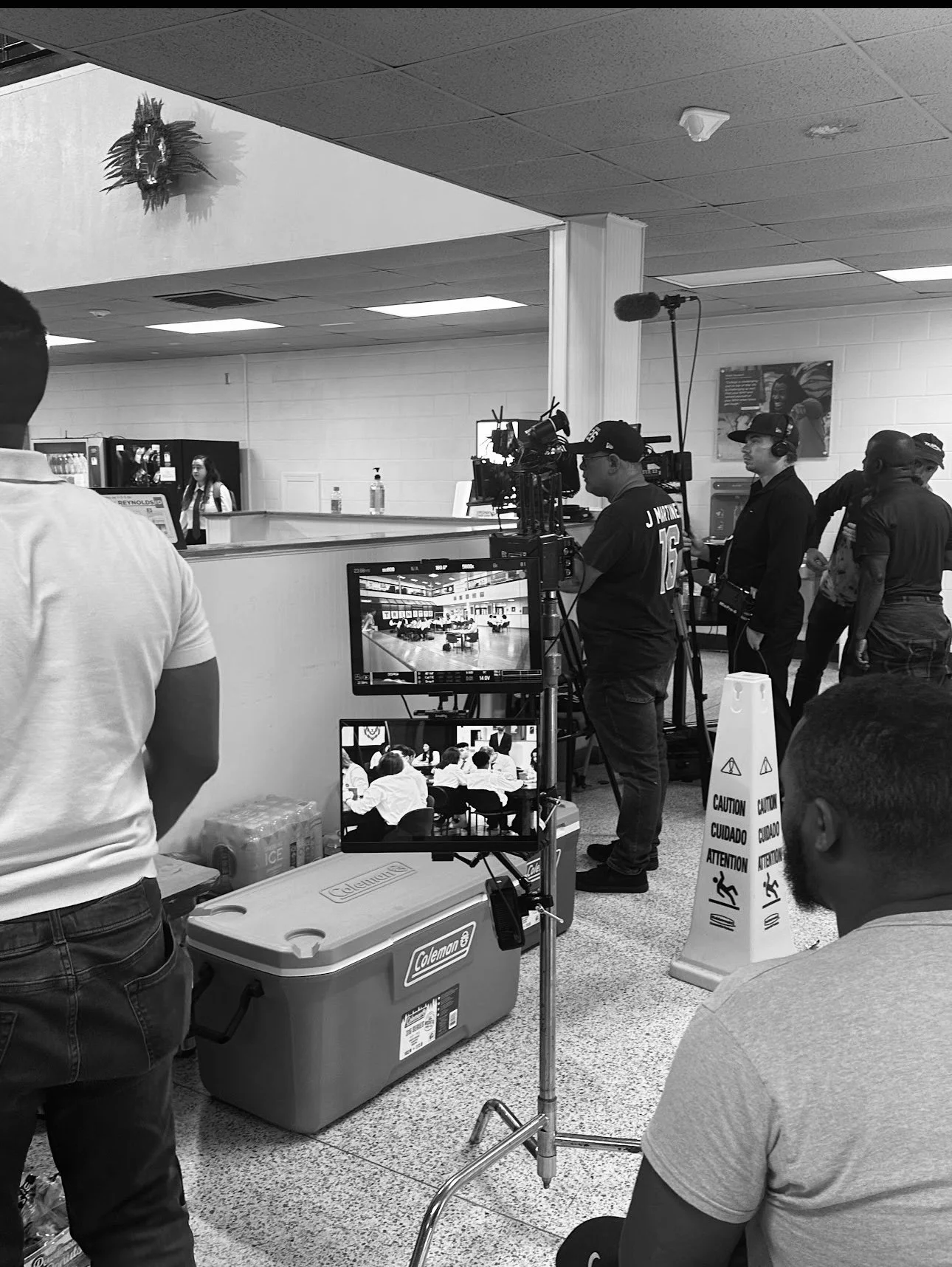 Filming crew operating camera equipment and monitors in a school hallway with students in the background and a caution sign on the floor.