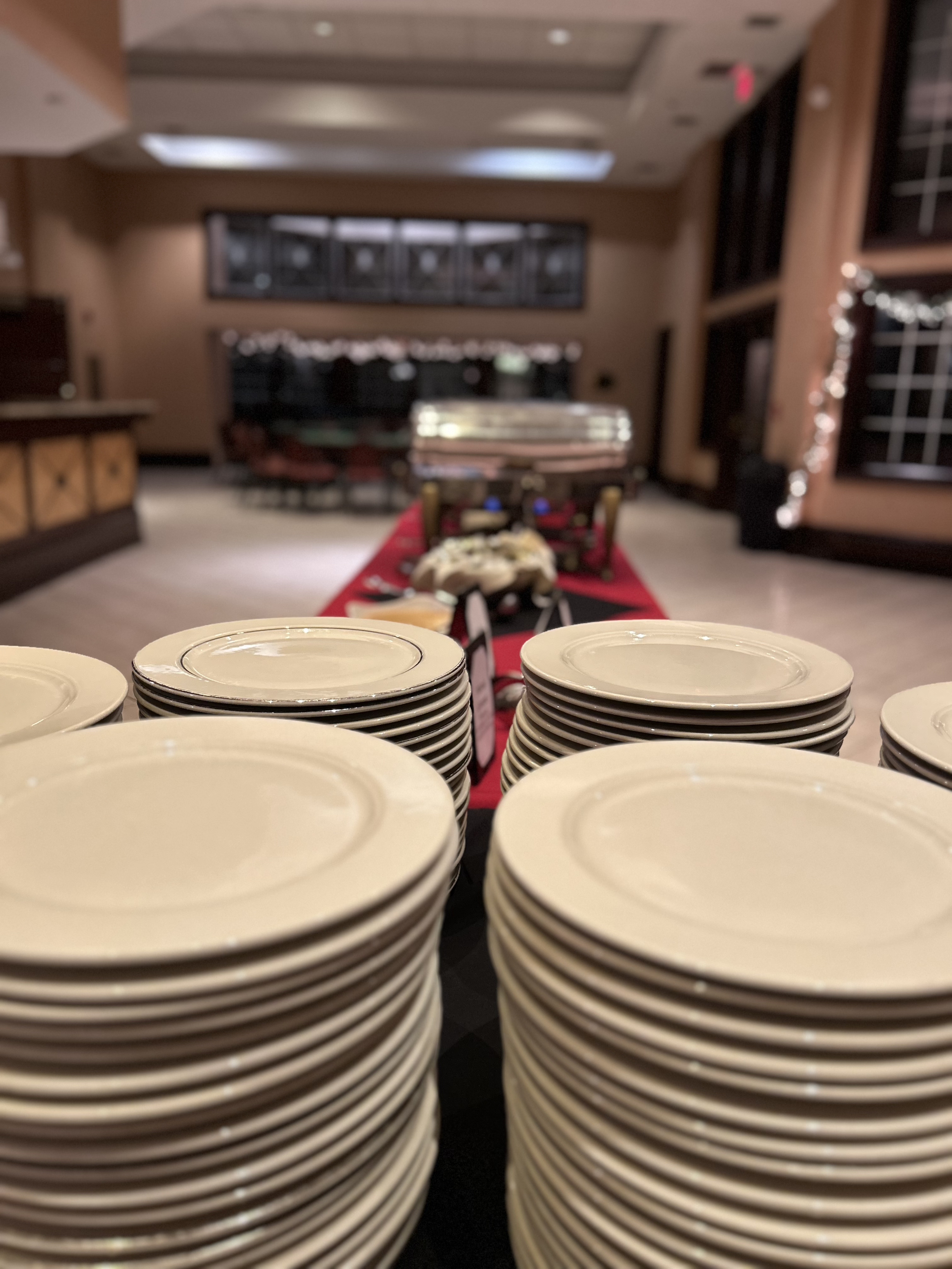 Multiple stacks of white plates arranged on a black table, set in a room with a red carpet, decorated for a celebration or event, with tables and chairs visible in the background.