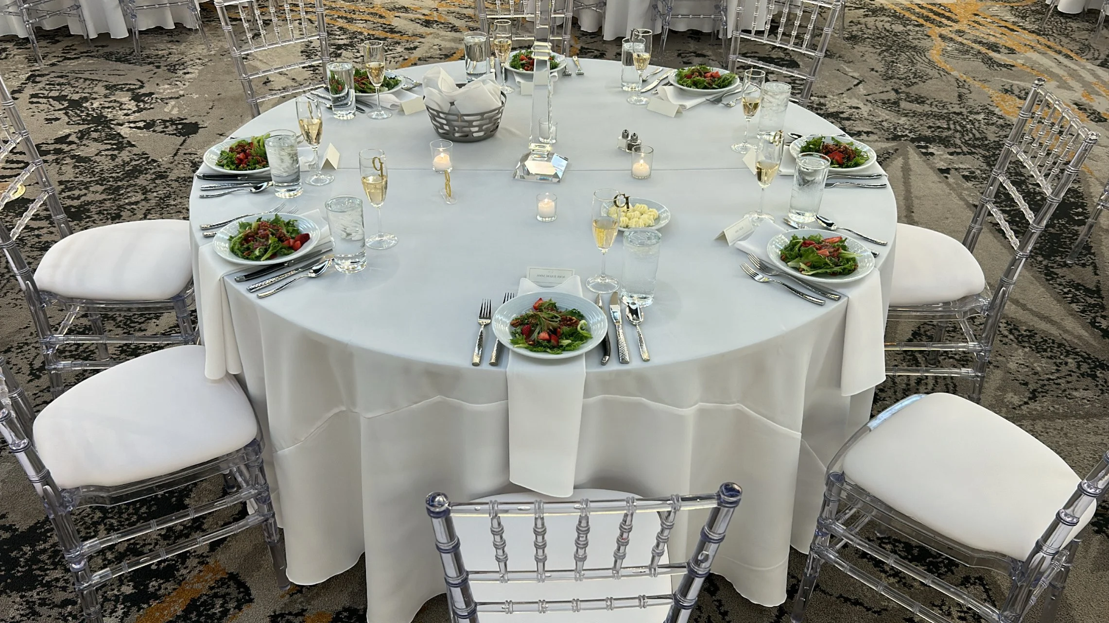 Round banquet table set with white tablecloth, salad plates, silverware, water glasses, champagne flutes, candles, and small name cards, surrounded by clear acrylic chairs with white cushions, on a patterned carpet.