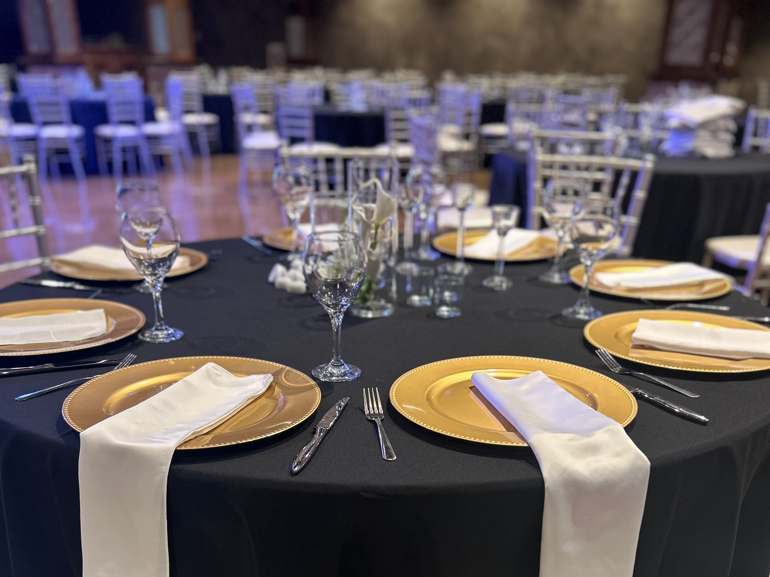 Round banquet table set for a formal event with gold chargers, white napkins, silverware, and water glasses, with a floral centerpiece and black tablecloths, in a venue with multiple similar tables and Chiavari chairs.