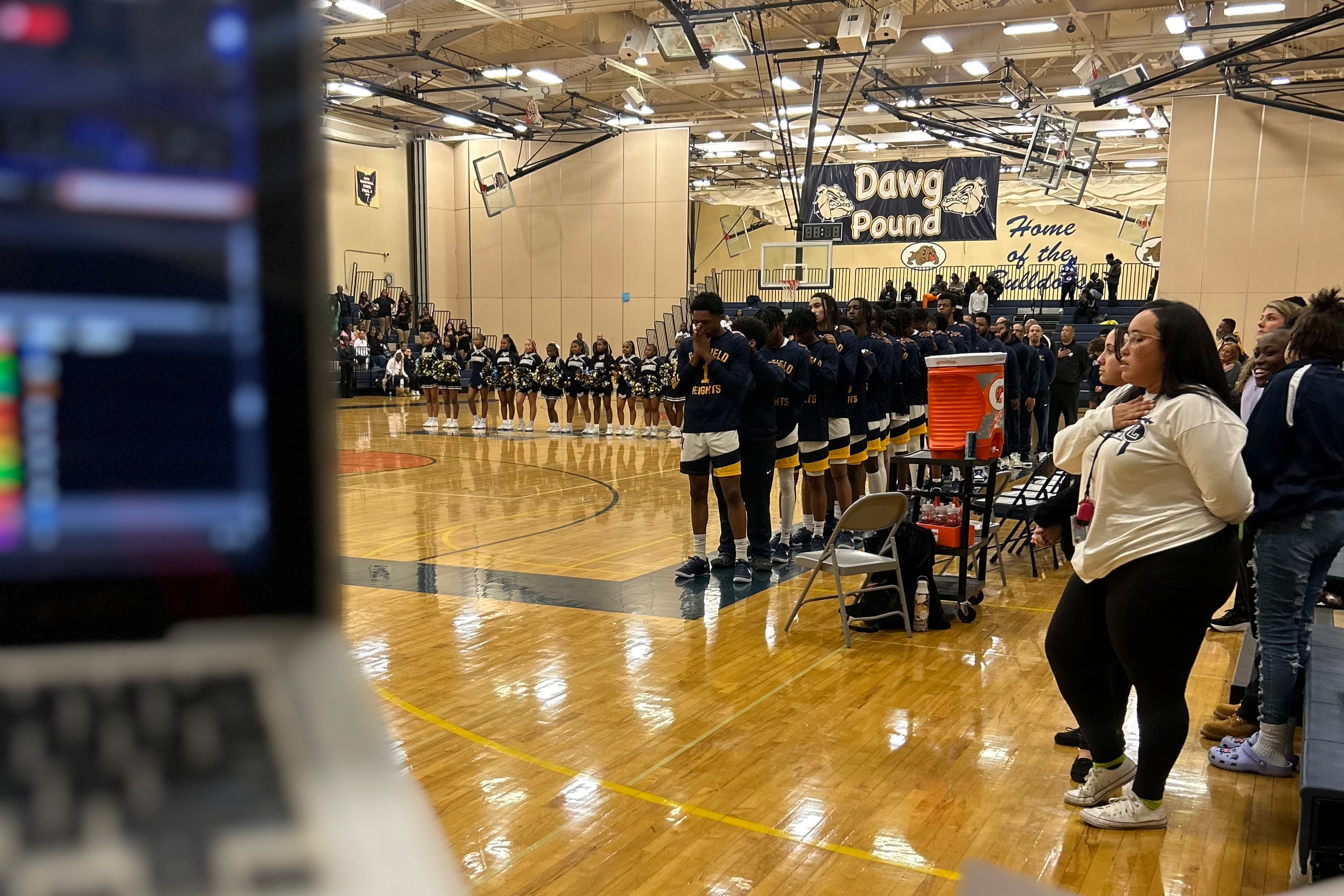 Basketball players and cheerleaders standing on a gym court during the national anthem, with spectators in the background, at a school game in a gymnasium.