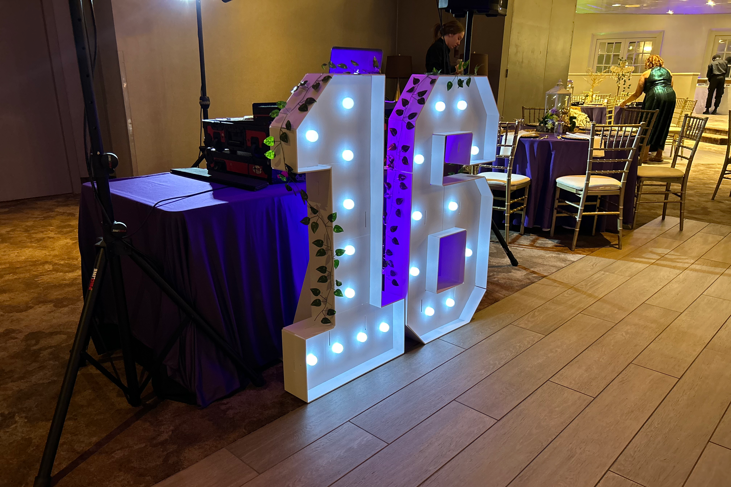 Large illuminated pink number 10 sign with decorative vines on each side, positioned on a wooden floor at an indoor event with purple tablecloths and gold chairs in the background.