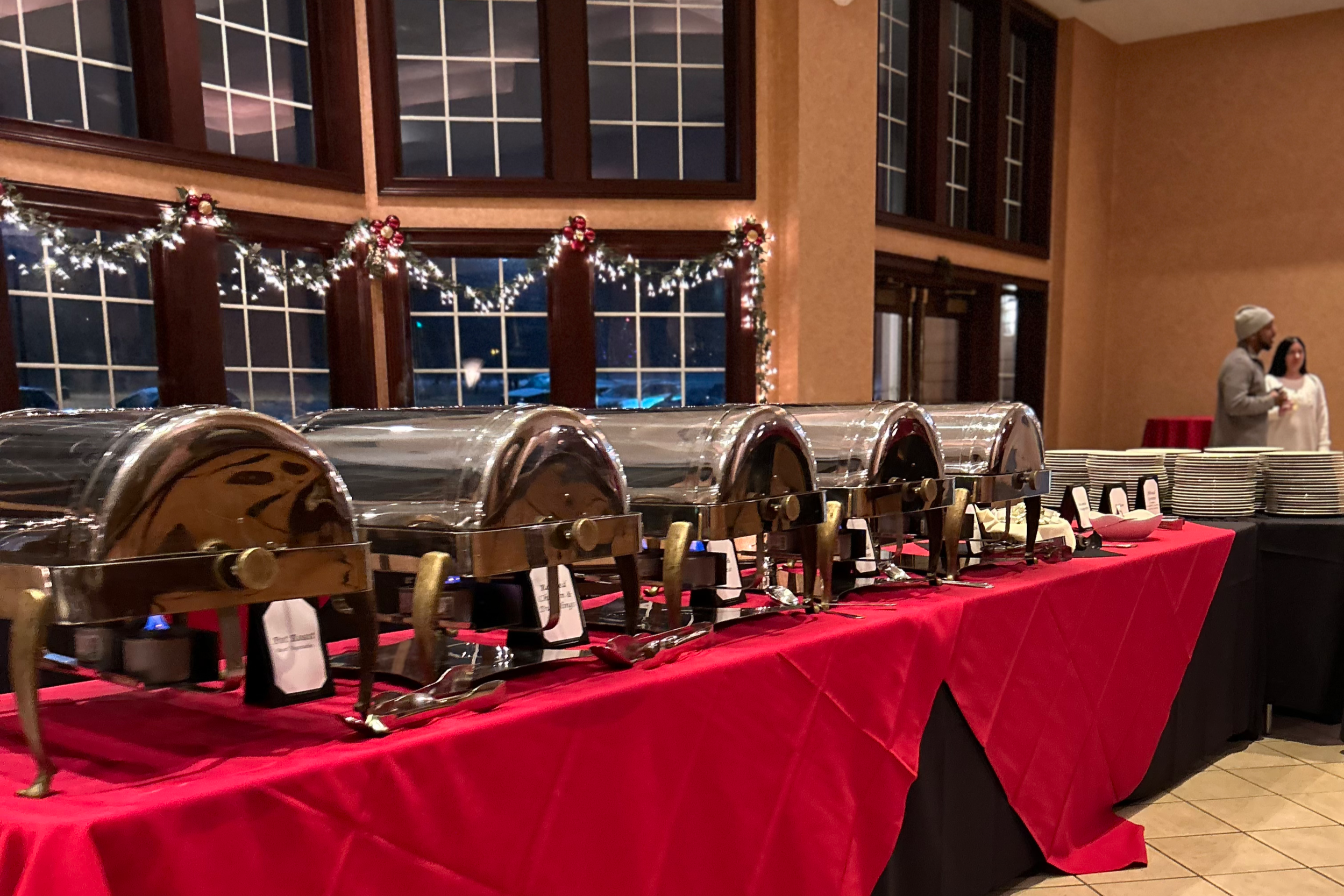 Buffet table with chafing dishes and plates at a holiday event, decorated with Christmas garland and ornaments.