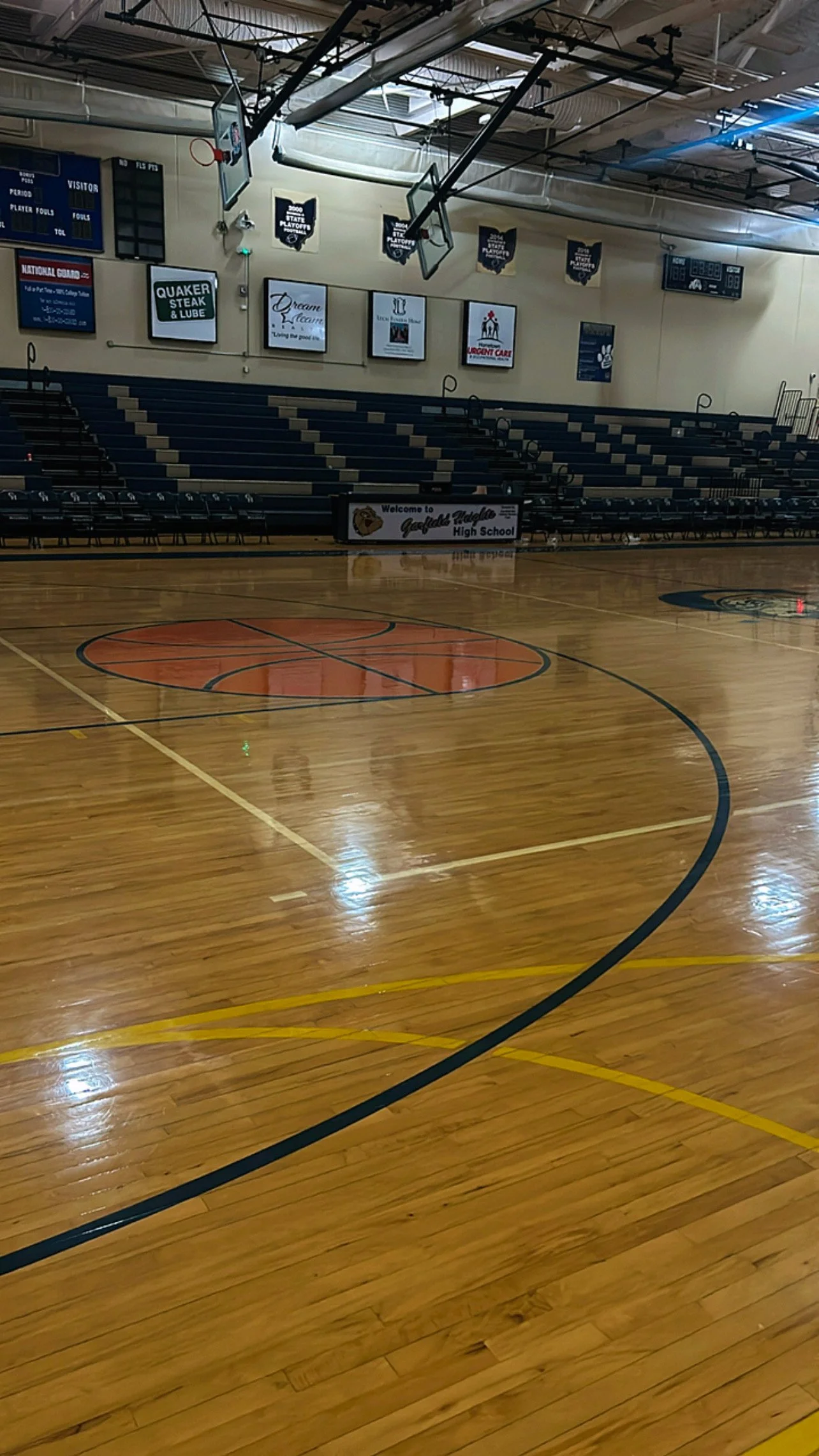 Empty indoor basketball court with a polished wooden floor, basketball hoops, and seating on one side. Banners and signs are visible on the wall behind the court, indicating it is Geddes High School.