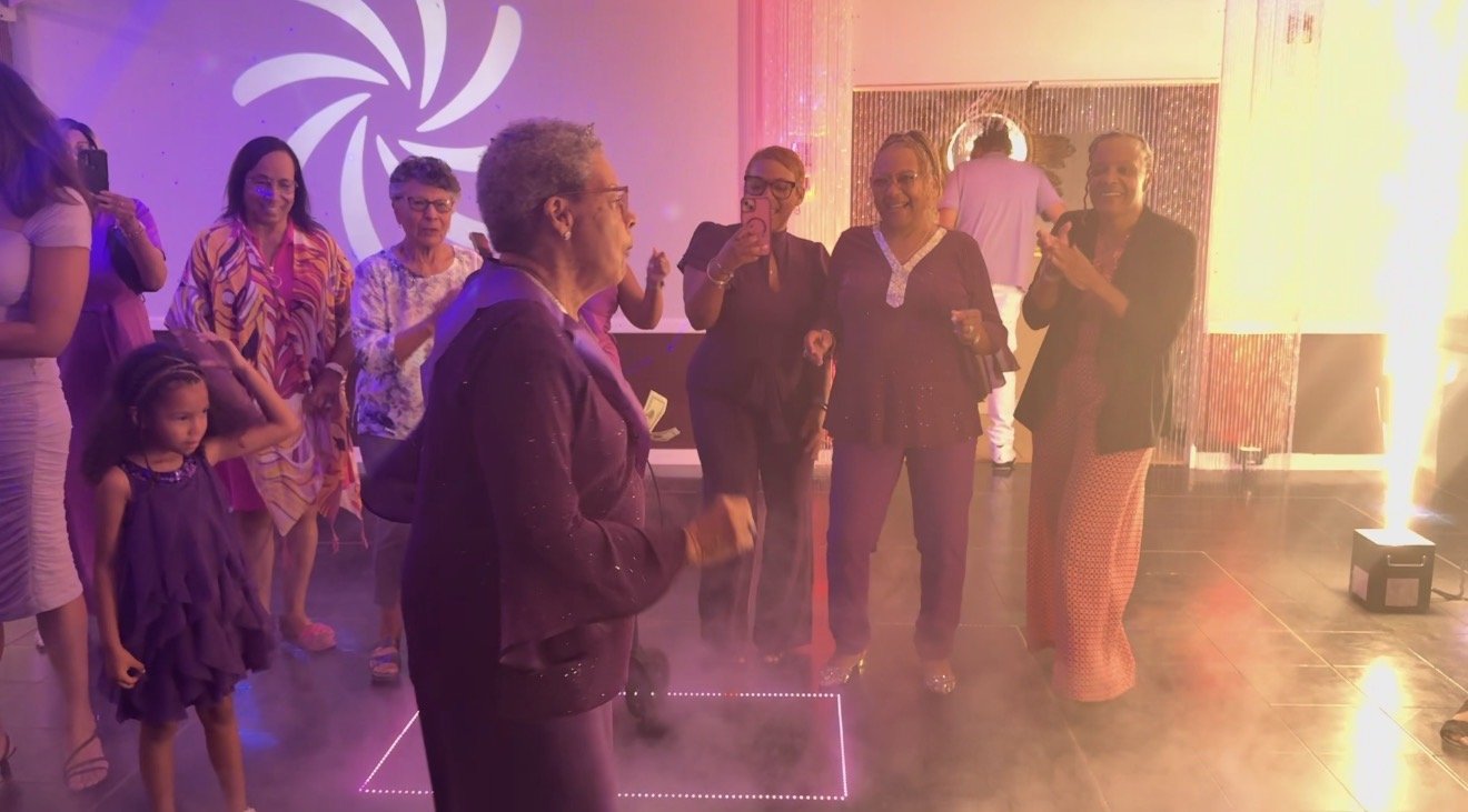 Group of women and a young girl dancing and socializing in a dimly lit party room with a colorful background and fog effects.