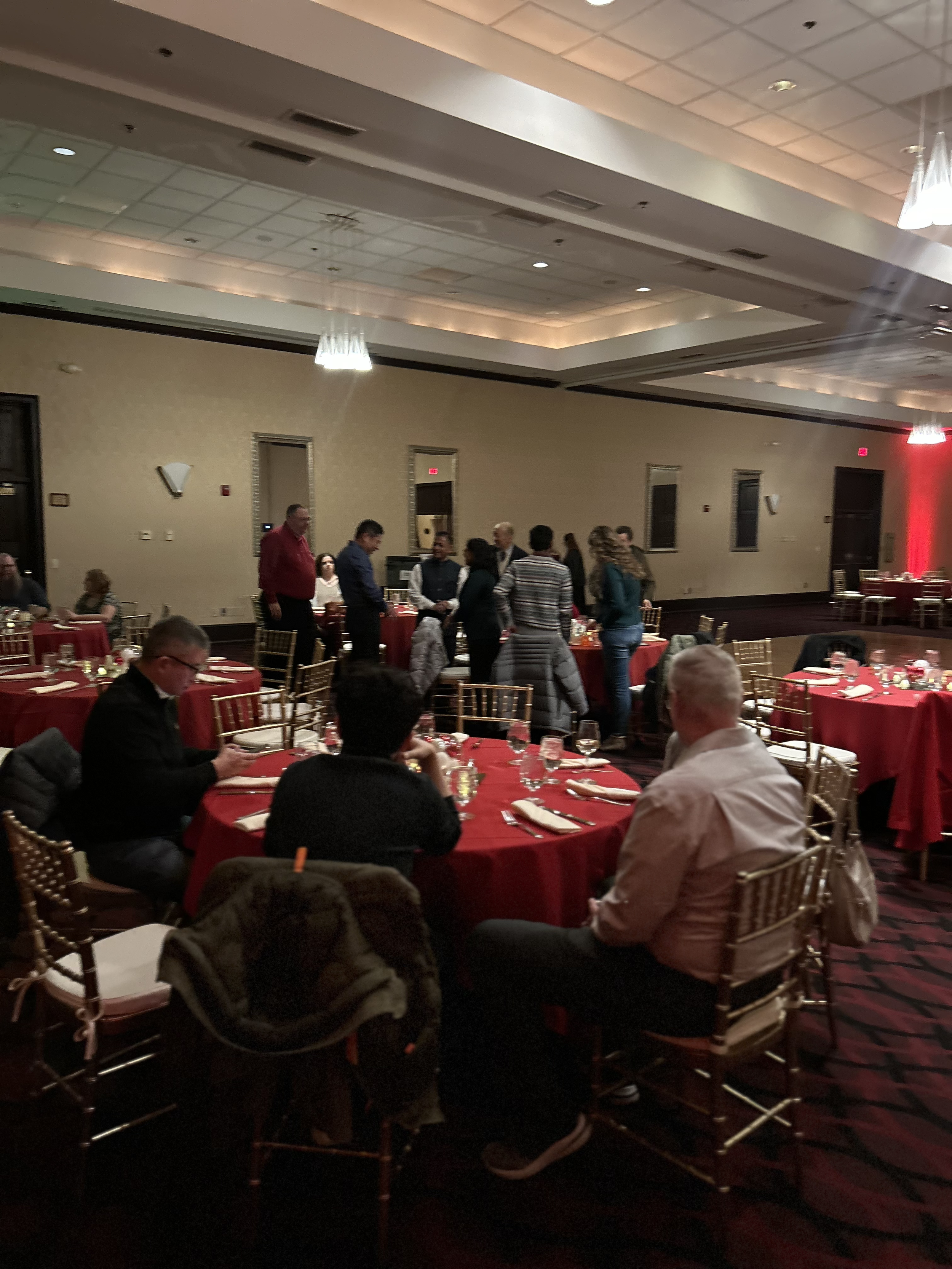 People dining in a banquet hall with round tables covered in red tablecloths, arranged for a formal event.