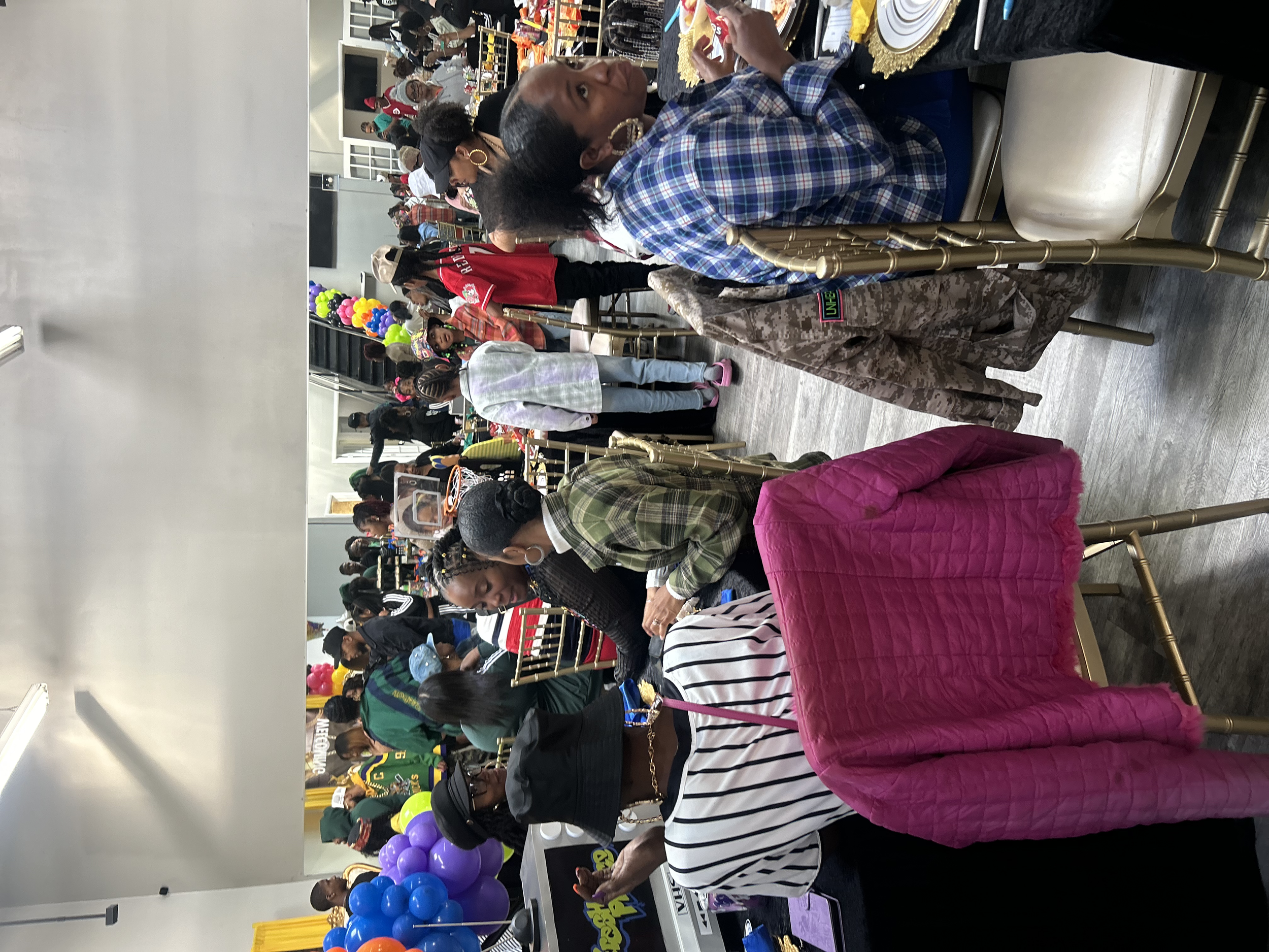 People gathered in a large indoor space, sitting at tables with colorful balloons and decorations, attending an event or celebration.