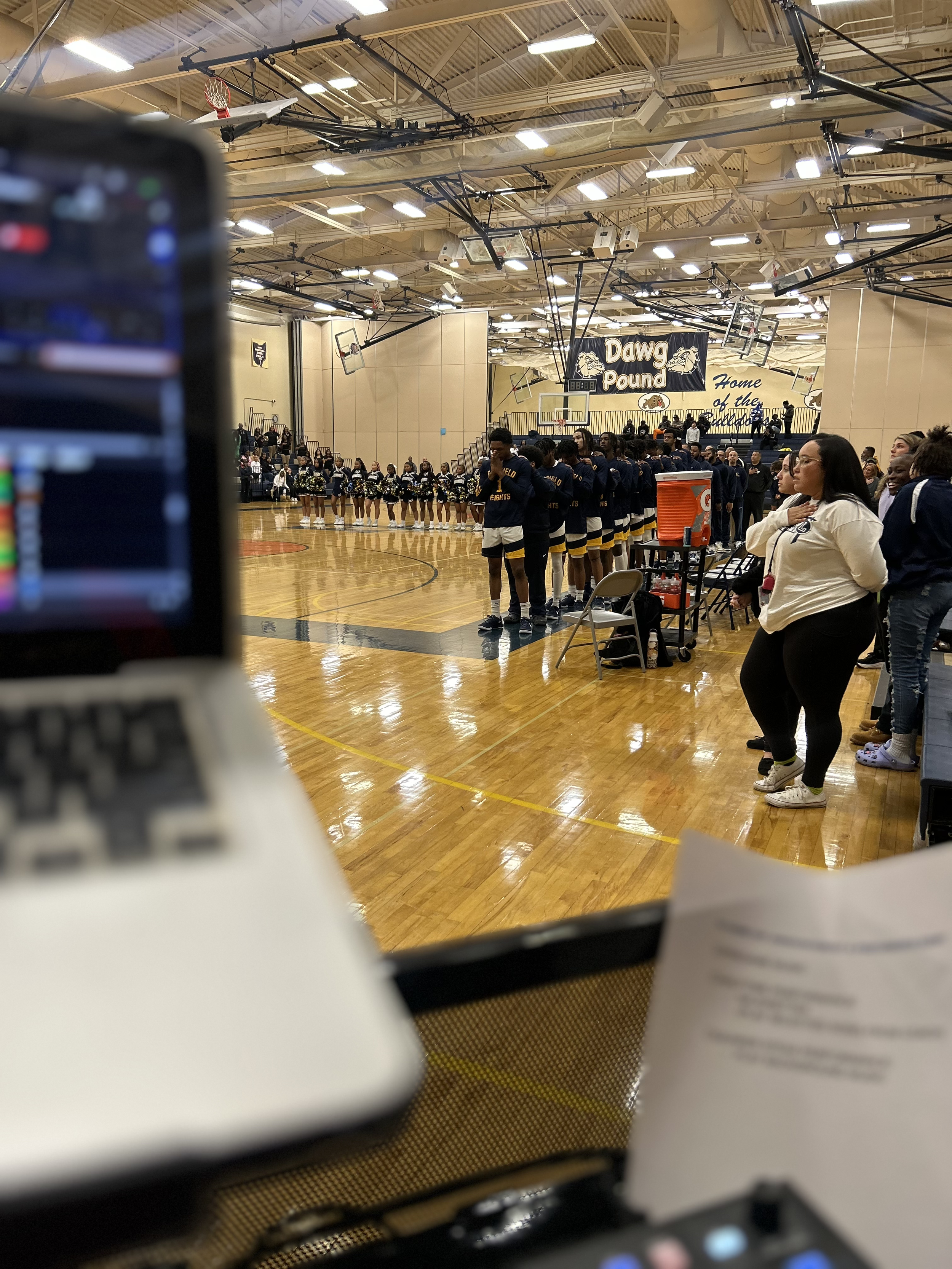A basketball game in a high school gymnasium with players standing in a line on the court and cheerleaders on the sidelines, with spectators seated in the bleachers. A sign reads 'Dawg Pound' at the back of the gym.