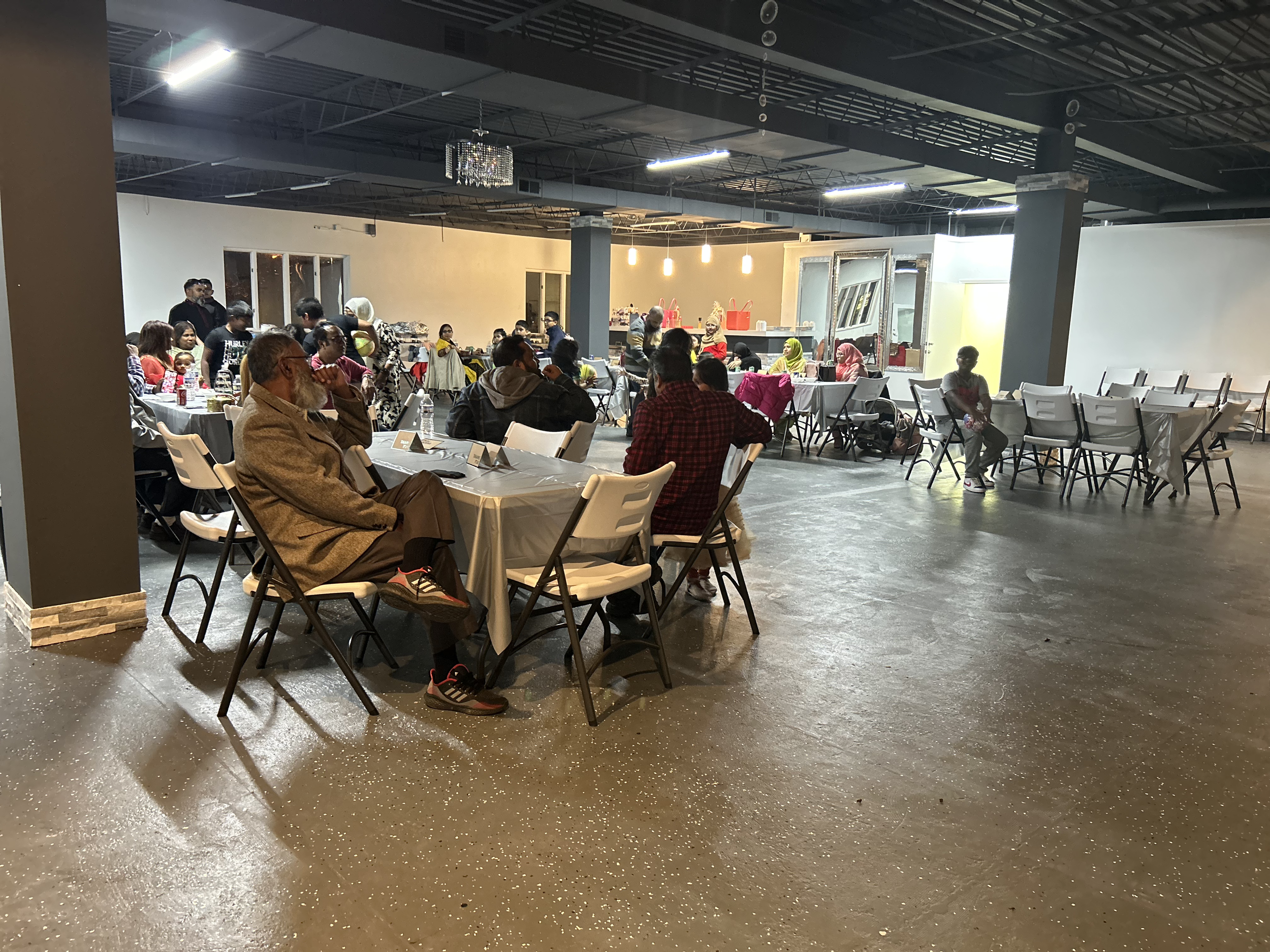 People dining and socializing in a spacious indoor event hall with tables, chairs, and a food truck in the background.