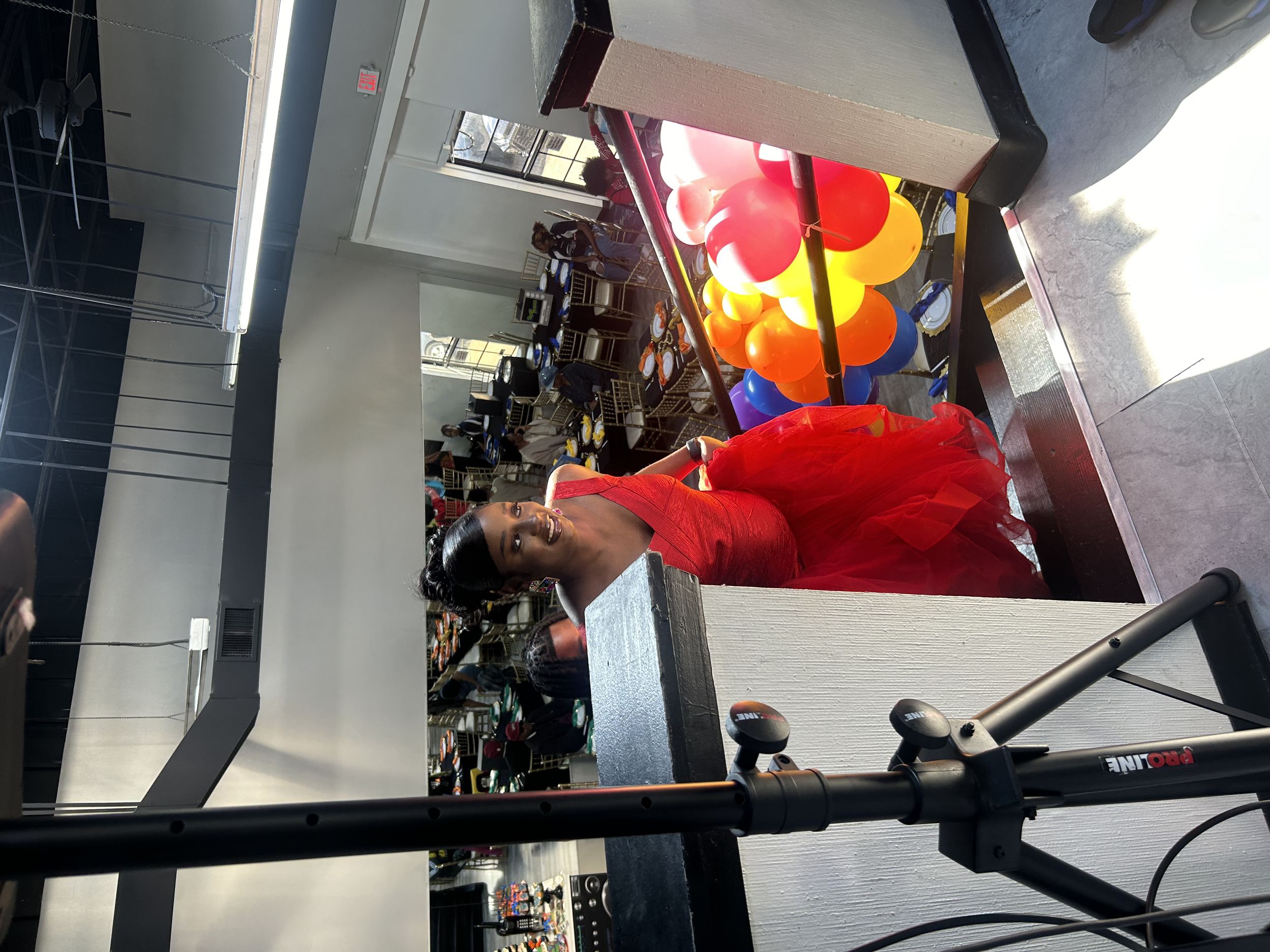 A woman in a red dress posing next to a colorful balloon display in a classroom or workshop setting with shelves of shoes and other items in the background.