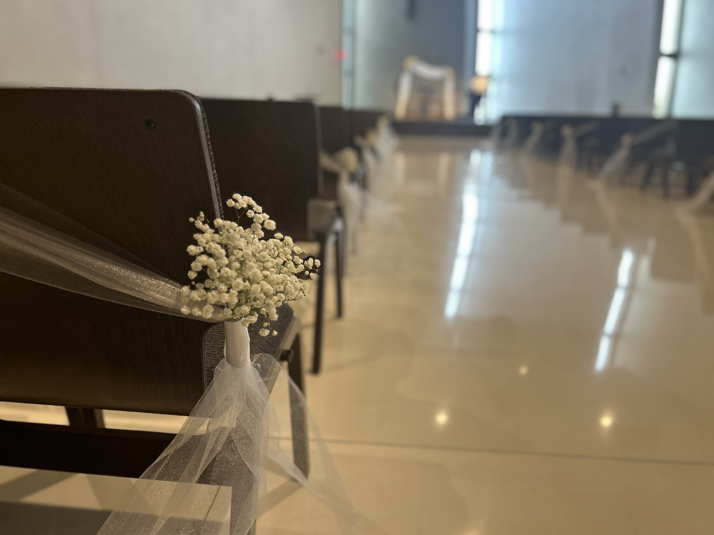 Row of empty dark wooden chairs decorated with small white flowers and sheer white ribbons, set up in a spacious, well-lit hall for an event.