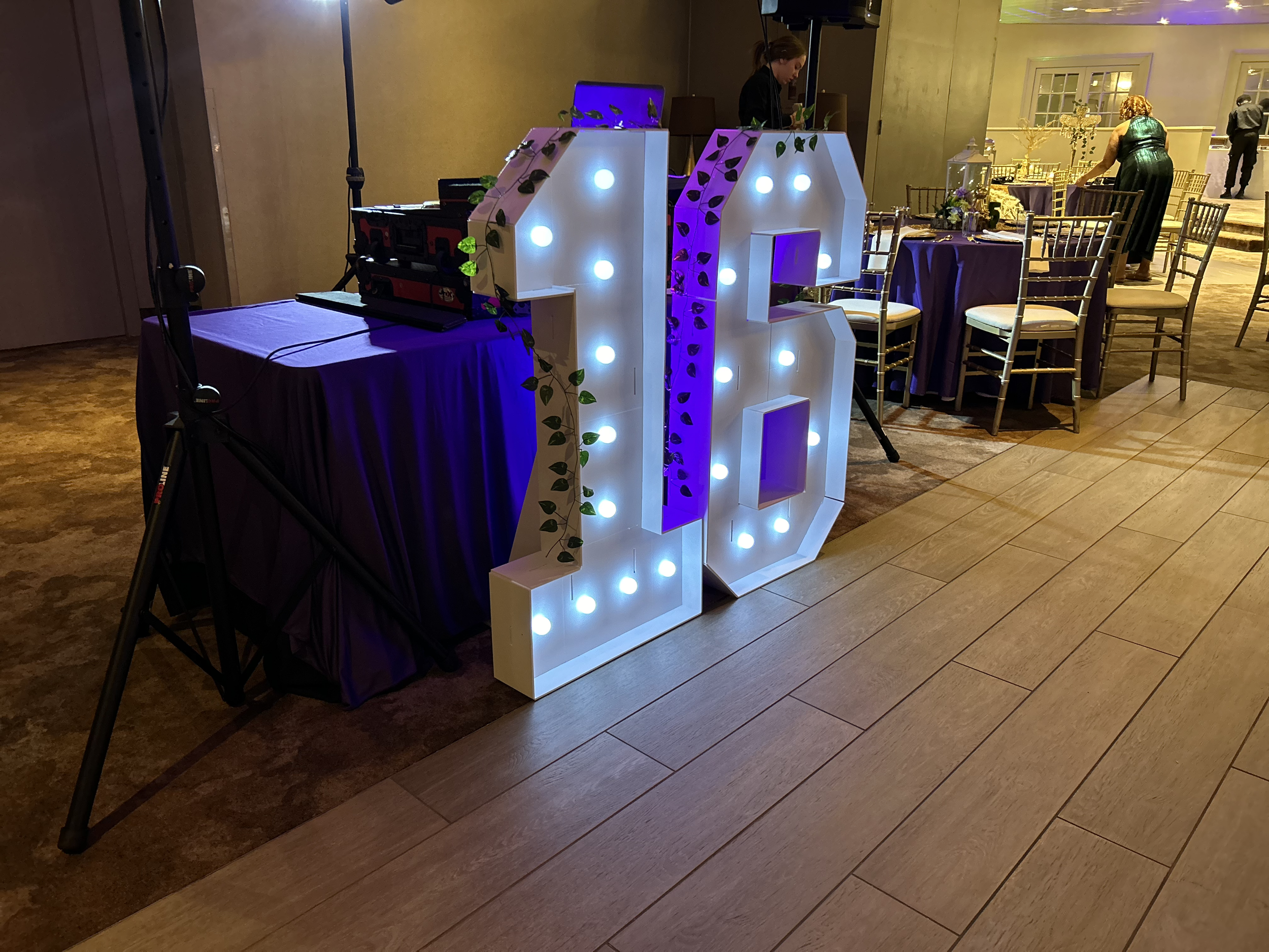 Large illuminated white decorative number 16 with purple accents and green vine decorations, set up at an event, with tables and chairs in the background.
