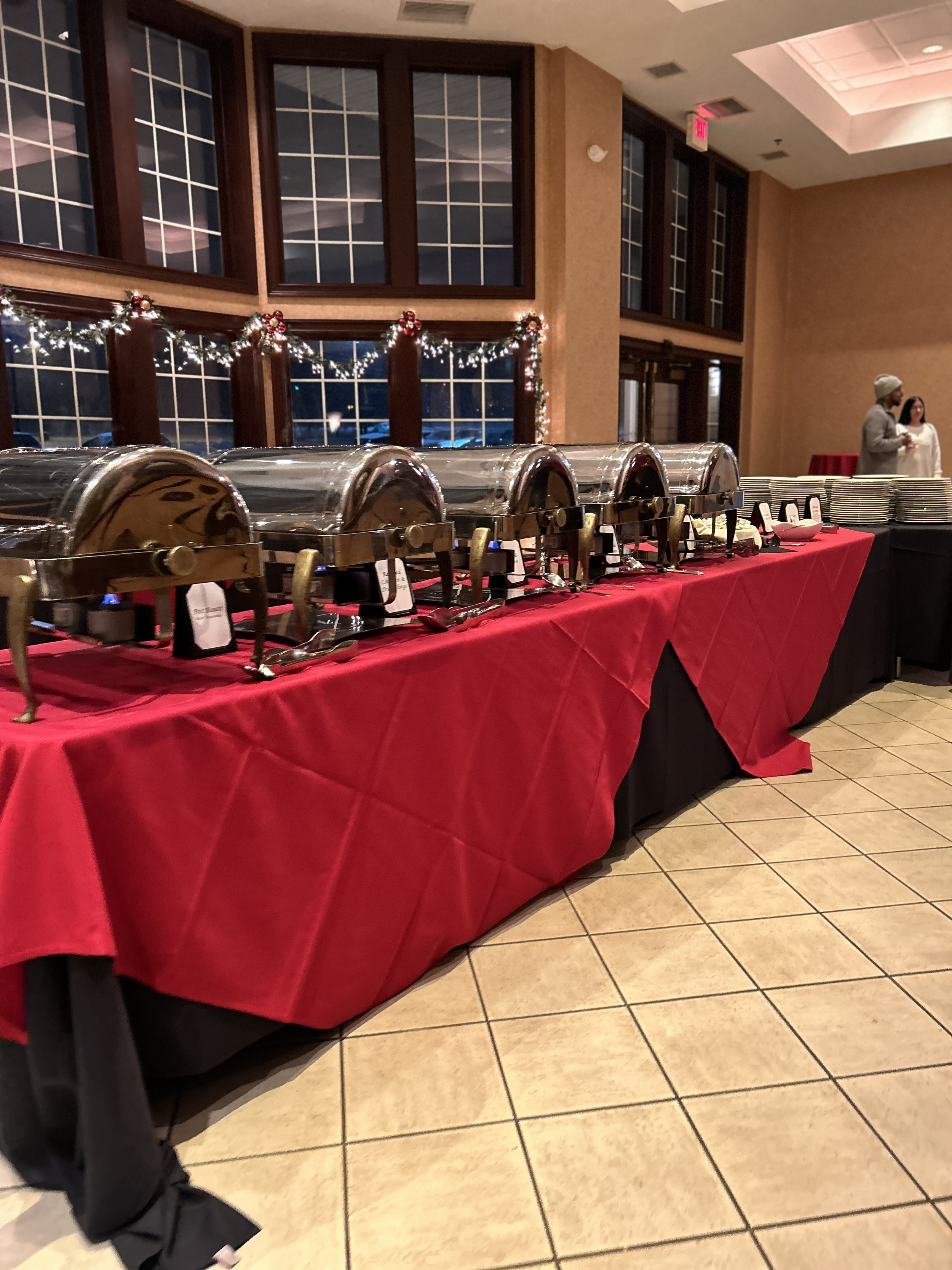 A buffet table decorated for Christmas with a red tablecloth, holding several chafing dishes, with plates and bowls on the side. In the background, a window with festive garland, and two people standing and talking near the table.