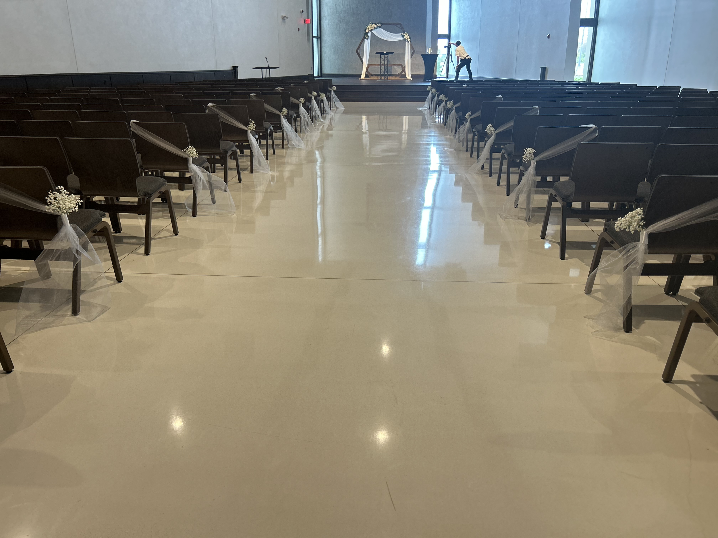 Empty wedding ceremony aisle decorated with white flowers and tulle, facing an altar with draped fabric and floral arrangements.