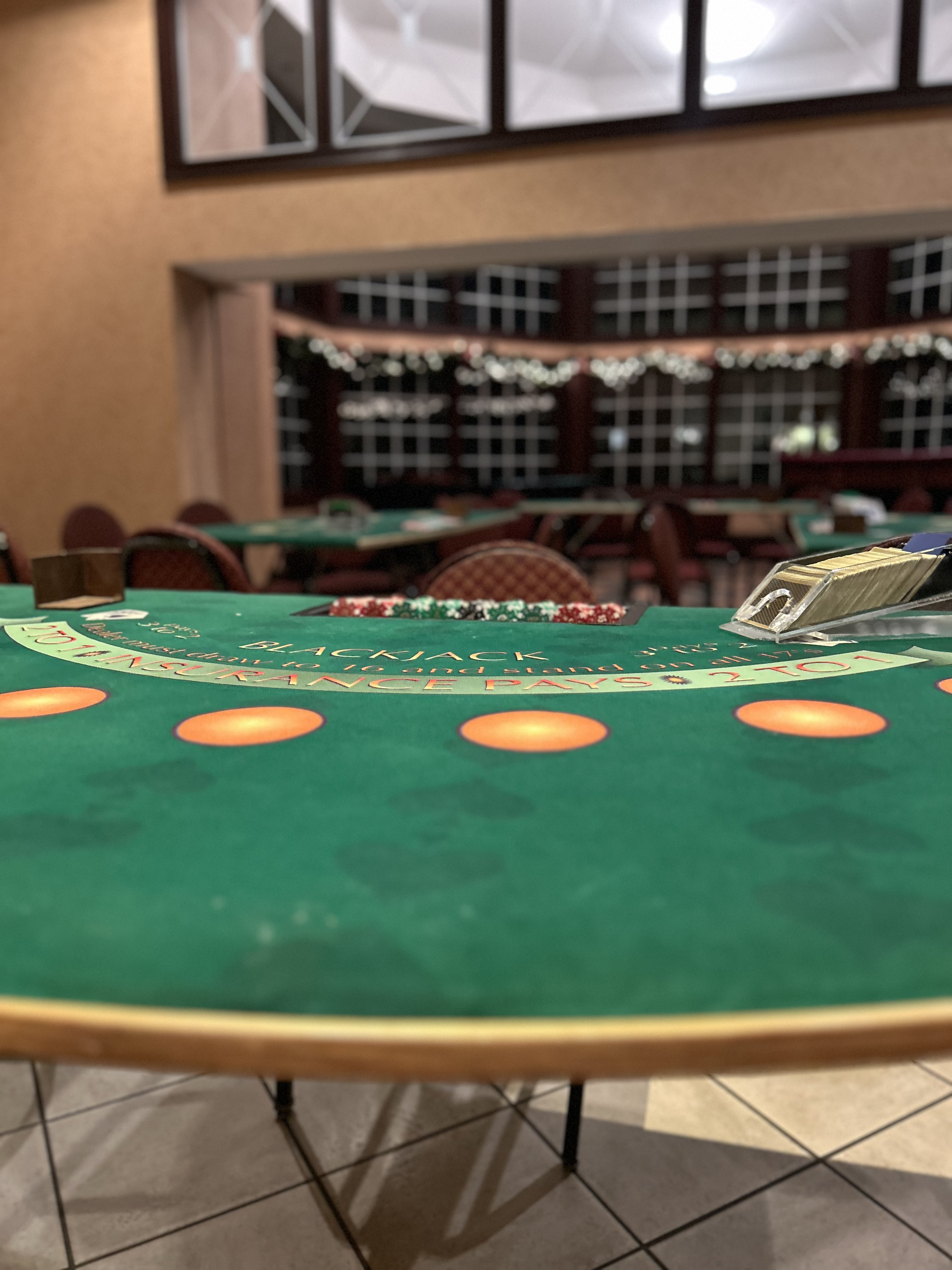 Close-up of a green blackjack table in a casino with chips and a dealer's shoe, with empty chairs and festive string lights in the background.