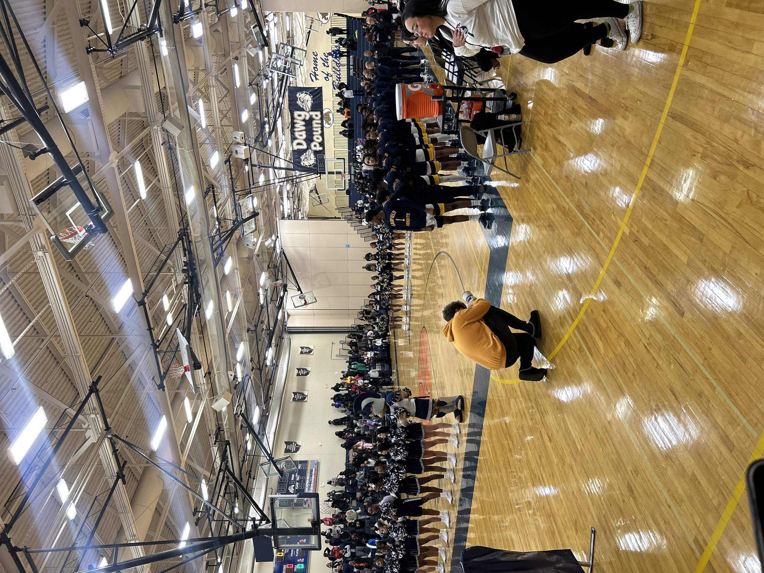 A basketball gymnasium filled with students, with some standing in line and others sitting against the wall. Two individuals are kneeling on the court, one in a yellow hoodie and the other near the students. The gym has wooden flooring, basketball ho