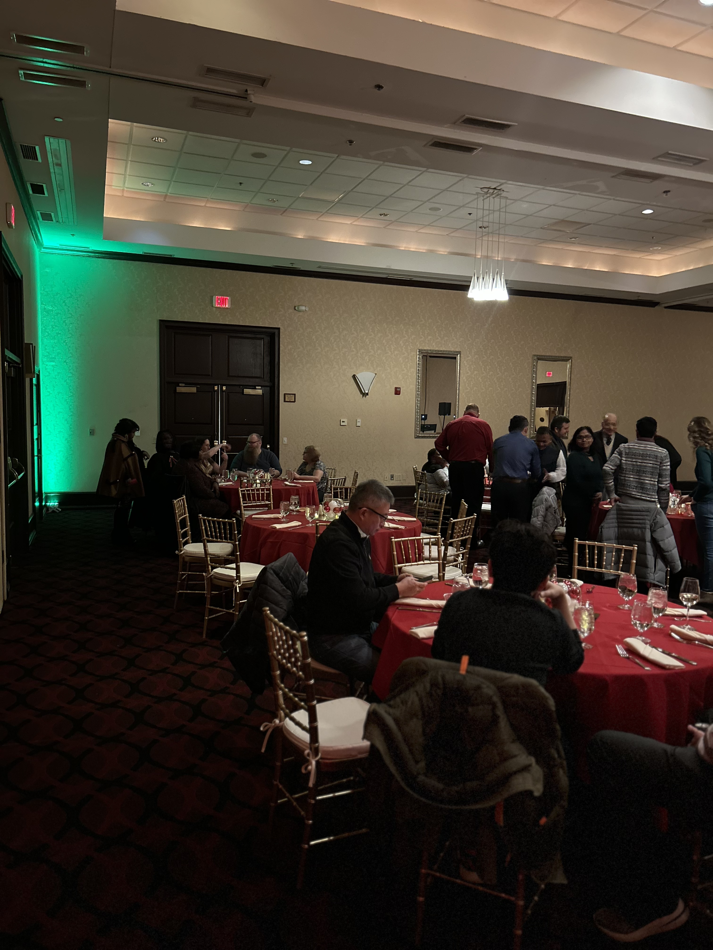 People seated at round tables with red tablecloths in a banquet hall, some standing and chatting, others seated and looking at their phones, with a green accent light on the left wall.