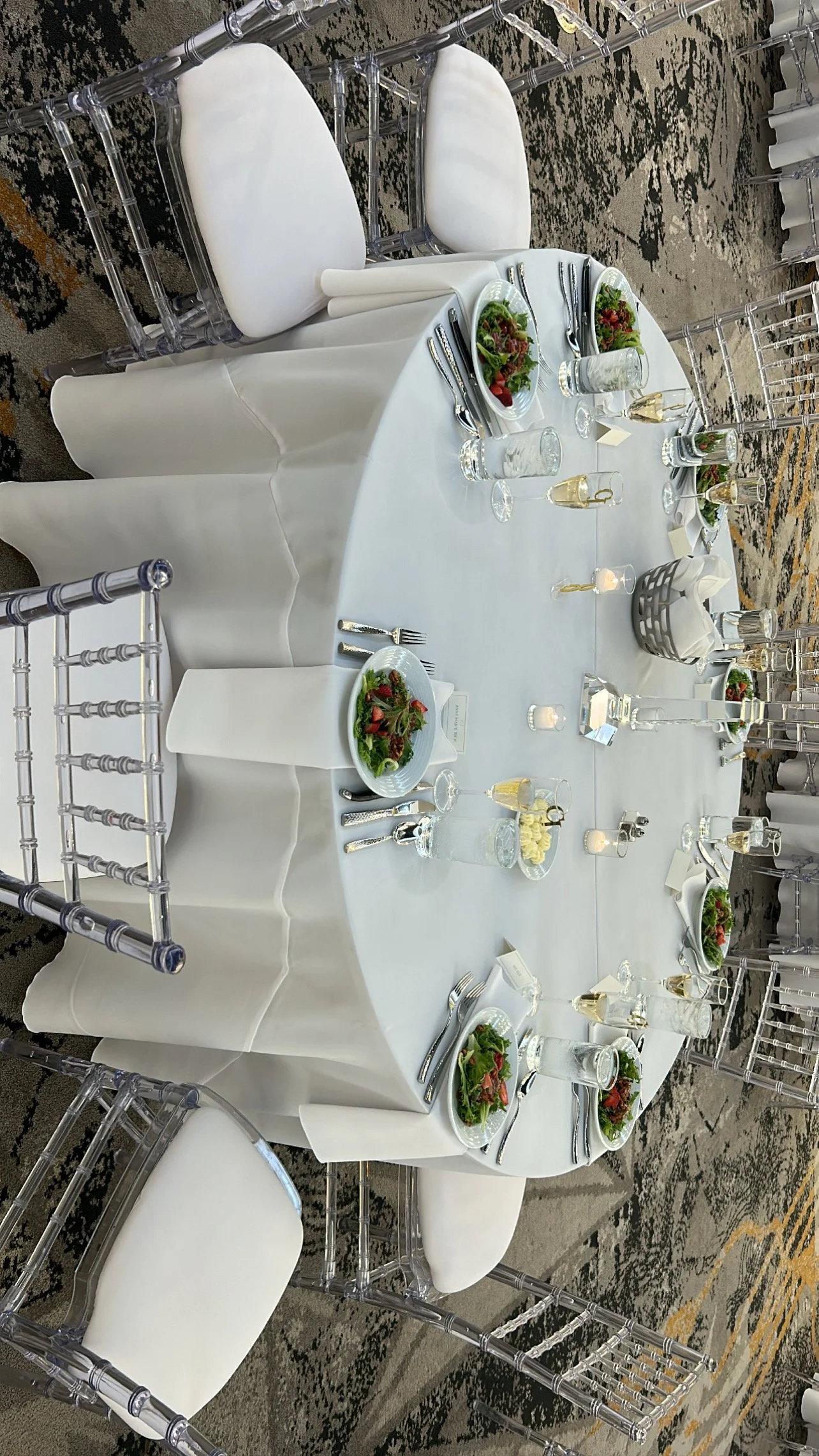 Round banquet table with white tablecloth, set for a meal with salad plates, silverware, water glasses, and small candles, surrounded by clear acrylic chairs