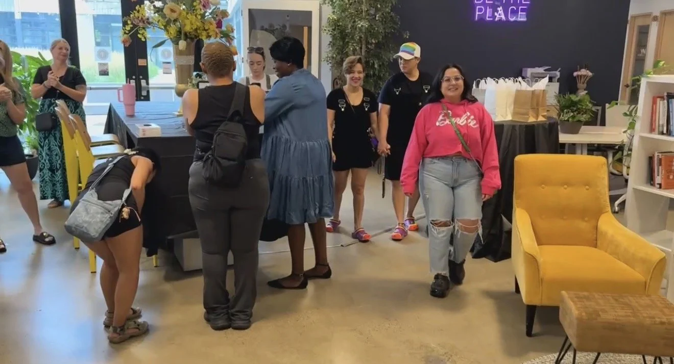 A group of people gathered in a modern indoor space, some are standing and chatting while others are walking or leaning over a table. The decor includes a large flower arrangement, a yellow armchair, and a bookshelf, with a neon sign that reads 'BE U