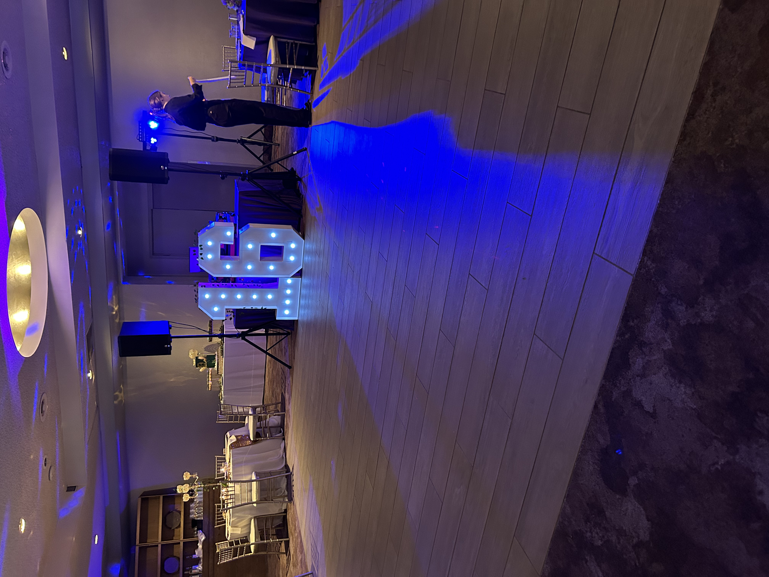 The image shows an event space with a wooden dance floor, blue lighting, and a large illuminated number 9 centerpiece. There is a person taking photos, a table with decorations and chairs, and a DJ setup in the background.