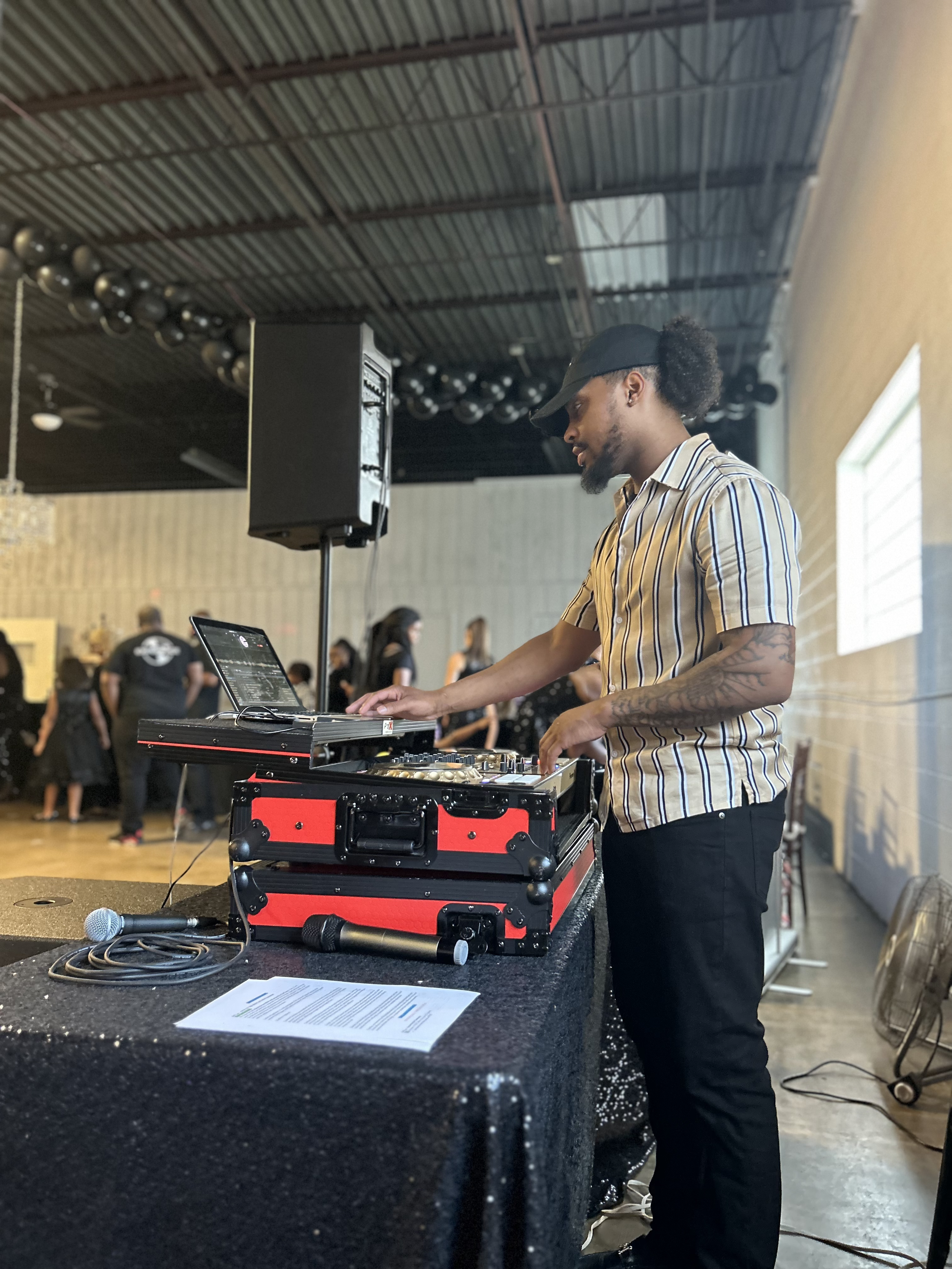 A man DJing at a live event, wearing a striped shirt and black cap, with a music mixer, laptop, and microphone on the table, in an industrial-style venue with other people in the background.