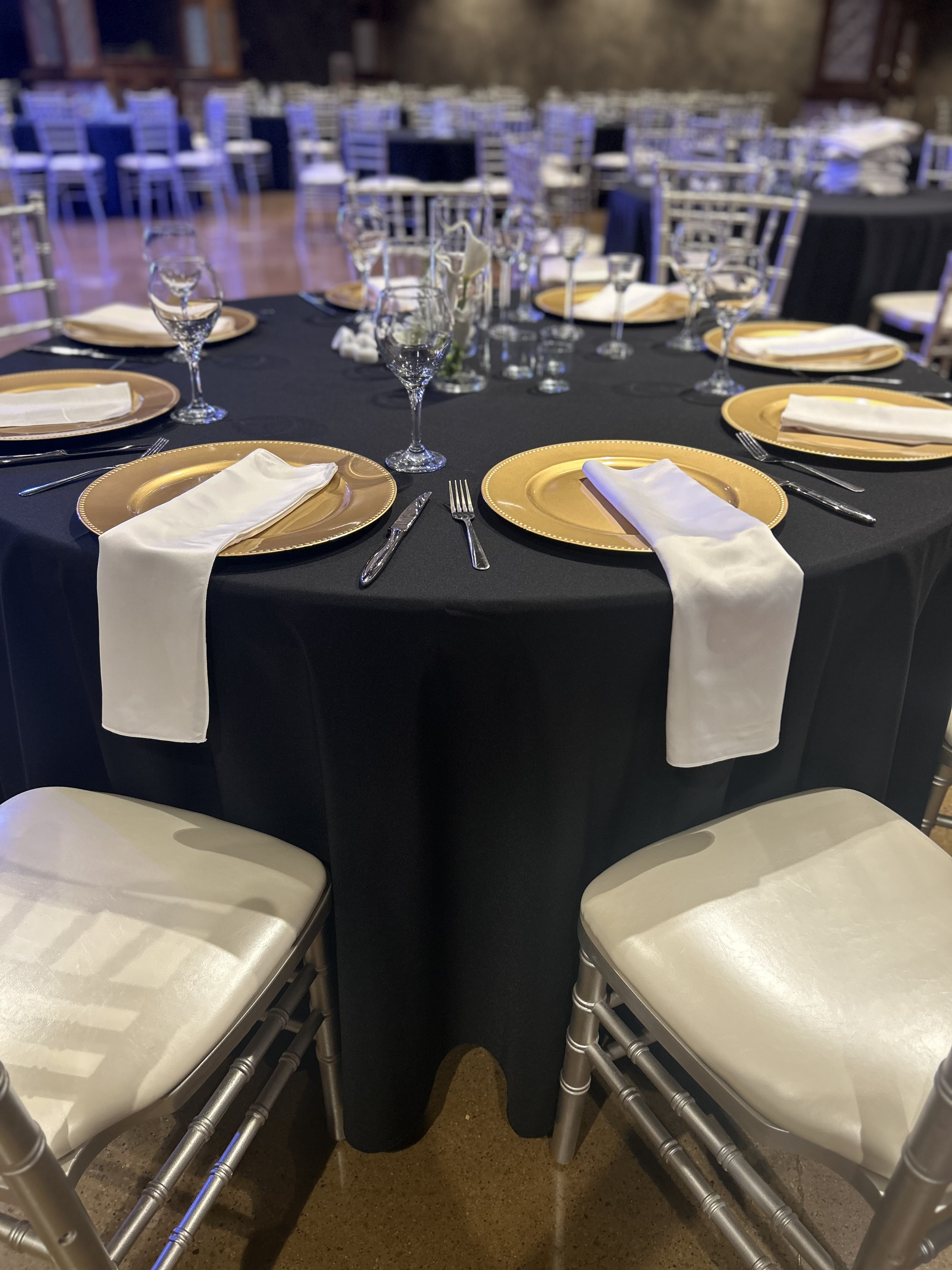 Round banquet table set for a formal event with black tablecloth, gold chargers, white napkins, and crystal glasses, surrounded by silver Chiavari chairs.