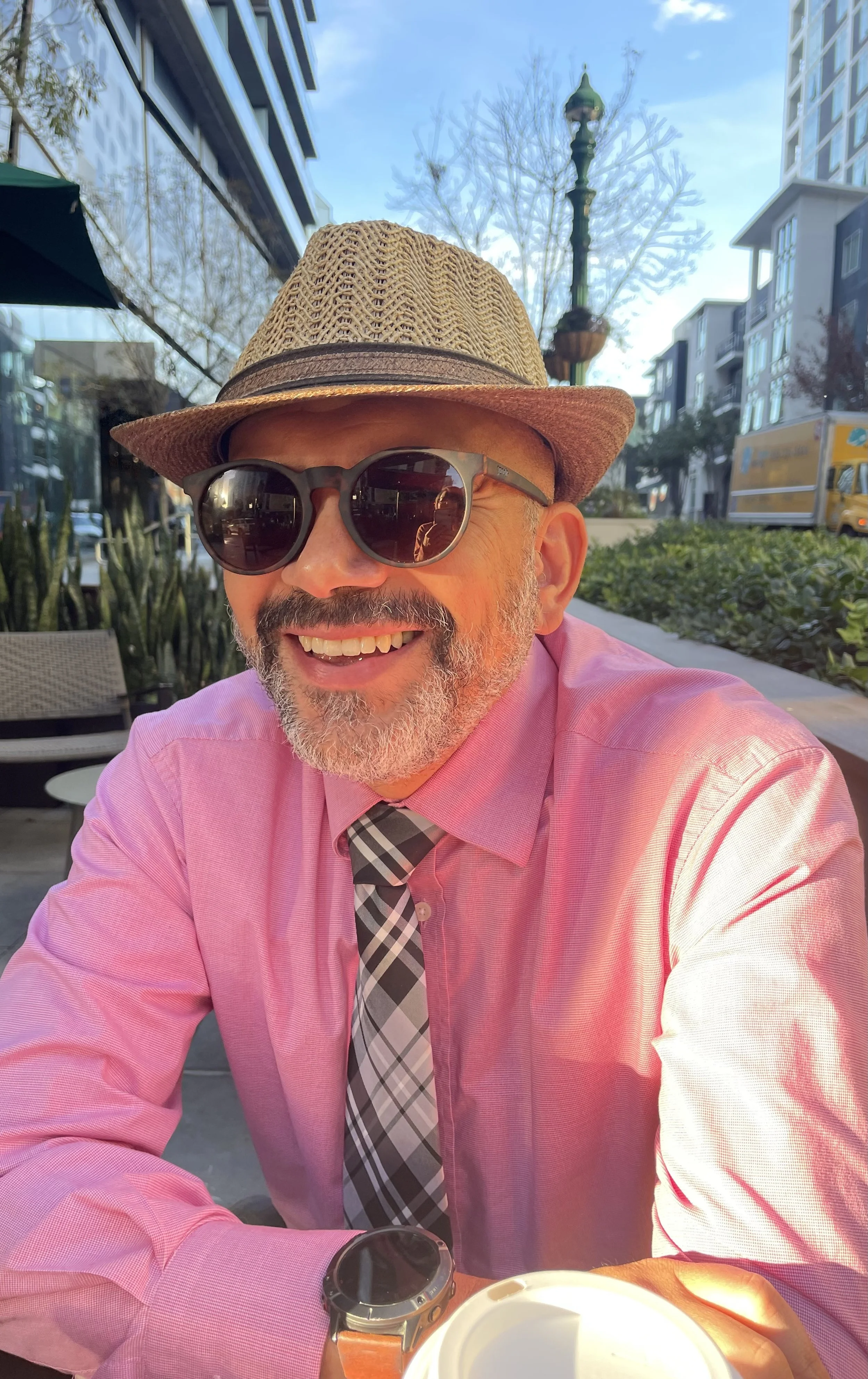 A man wearing a straw fedora, dark sunglasses, a pink dress shirt, a plaid tie, and a smartwatch, smiling while sitting outdoors at a table with a paper coffee cup in front of him, in an urban area with modern buildings, a streetlamp, and leafless trees in the background.