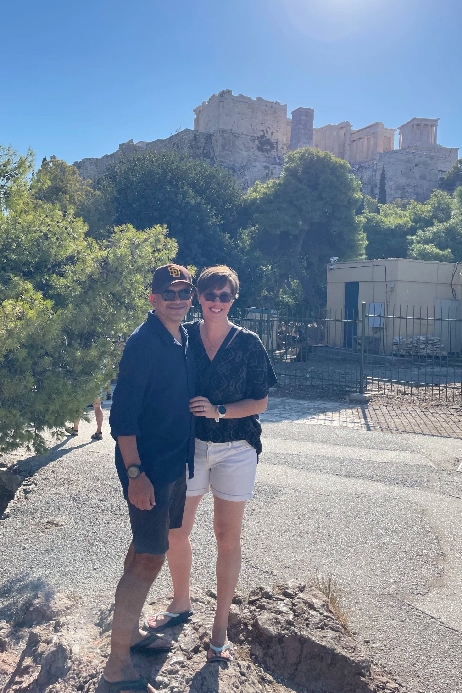 A couple standing outdoors near the Acropolis in Athens, Greece. The man is wearing a baseball cap, sunglasses, and casual clothing. The woman is smiling, wearing a patterned black shirt, white shorts, and sunglasses, with her arm around the man.