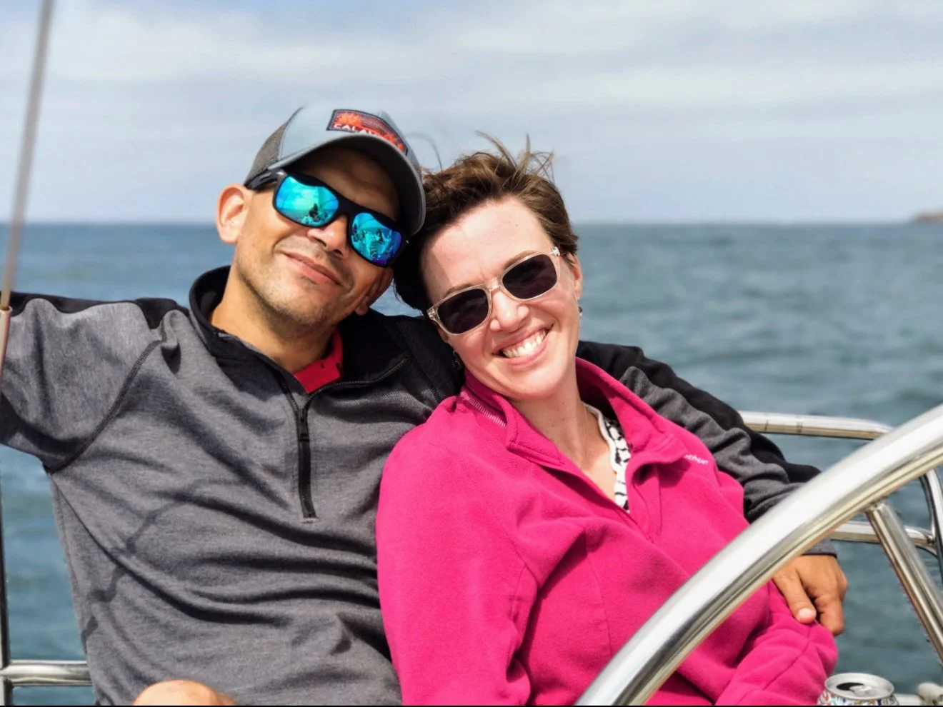 A smiling couple wearing sunglasses enjoying an afternoon of sailing in San Diego Harbor.  The man is wearing a cap and sunglasses and the woman has sunglasses and a pink pullover.