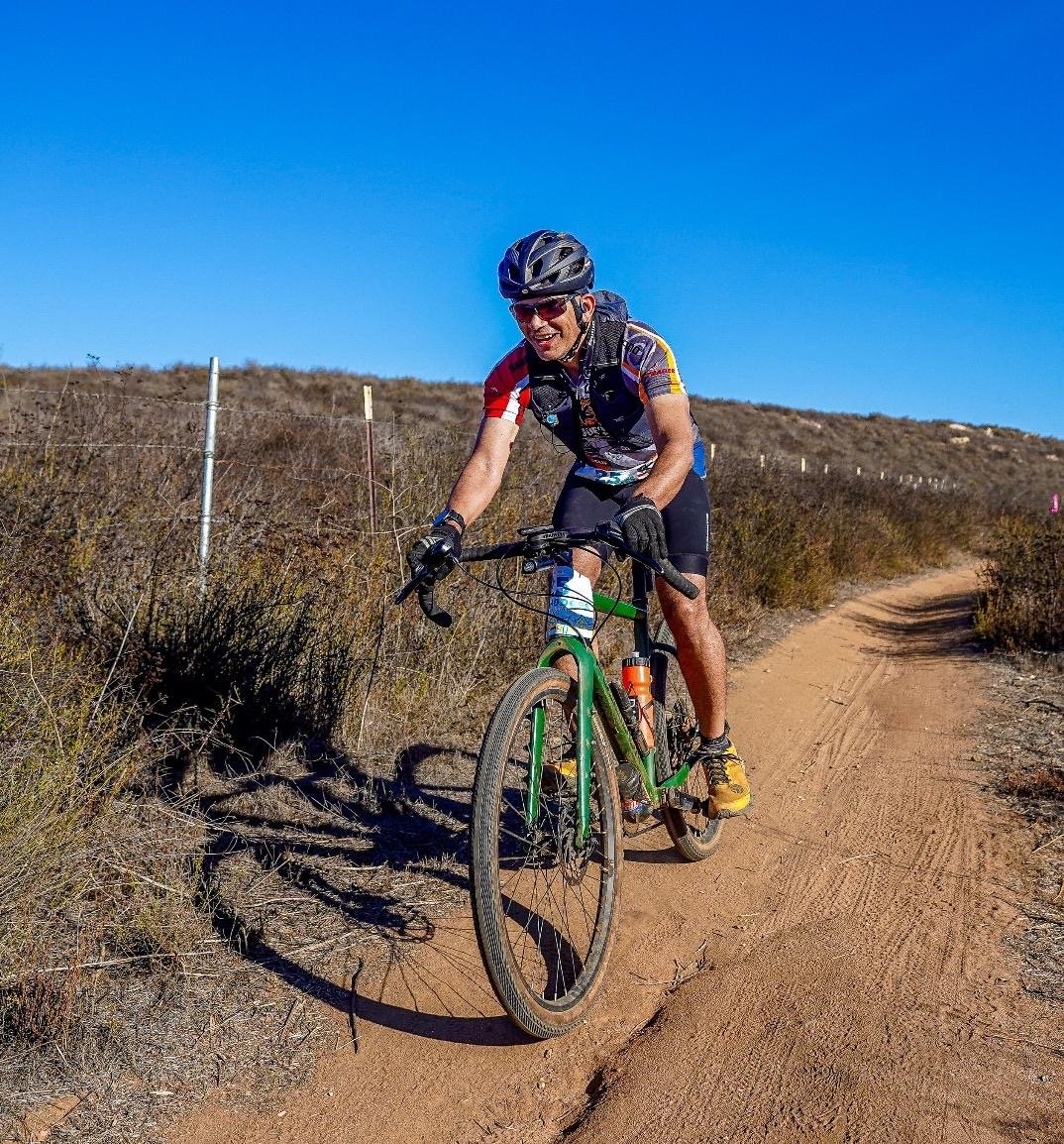 Man riding a mountain bike on a dirt trail in the dry, hilly terrain of Del Dios near Lake Hodges under a clear blue sky.