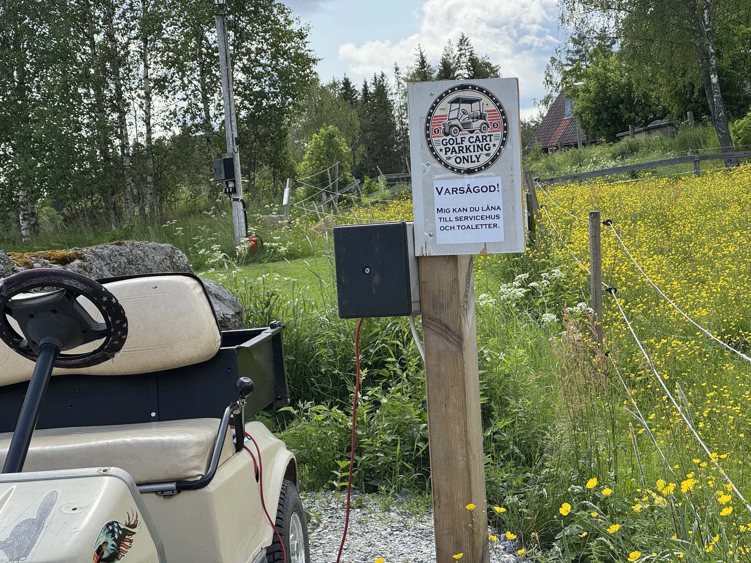 Skylt som anger att golfbilen kan lånas till servicehus och toaletter. Golfbil står bredvid skylten i ett grönområde med blommor.