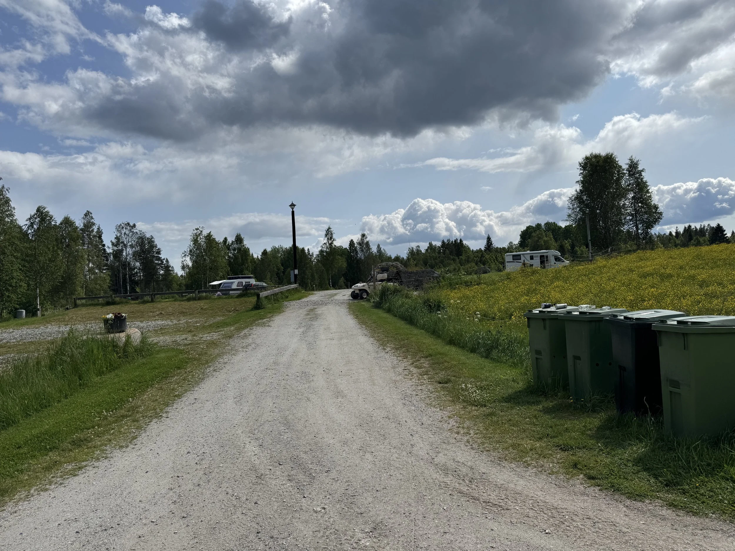 En väg med gräs och blommor på sidorna, omgiven av träd, med några husvagnar och bilar i bakgrunden mot en molnig himmel.