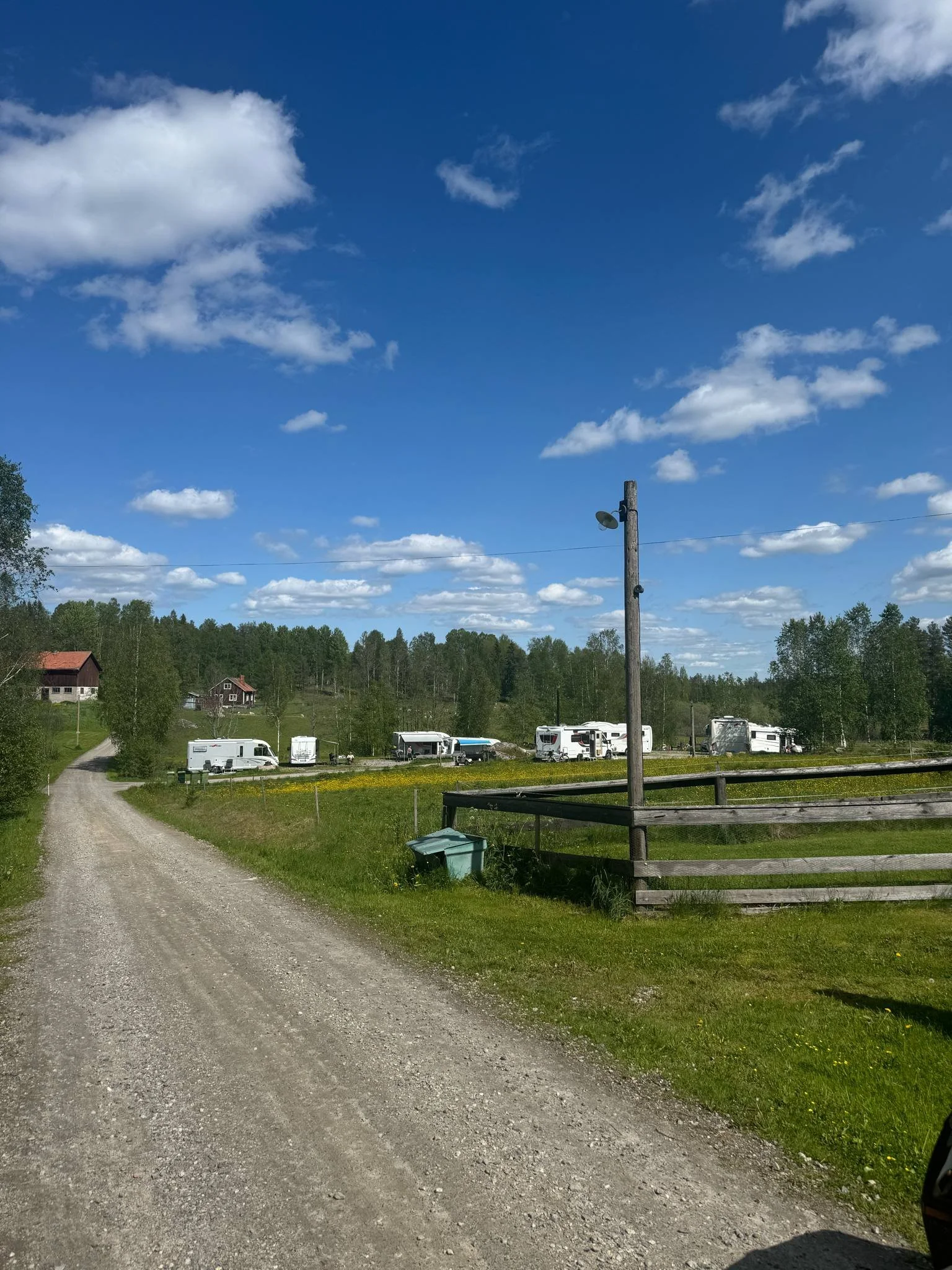 En grusväg i ett landskap med gröna träd och några hus i bakgrunden, samt flera husvagnar parkerade i ett fält, mot en blå himmel med spridda vita moln.