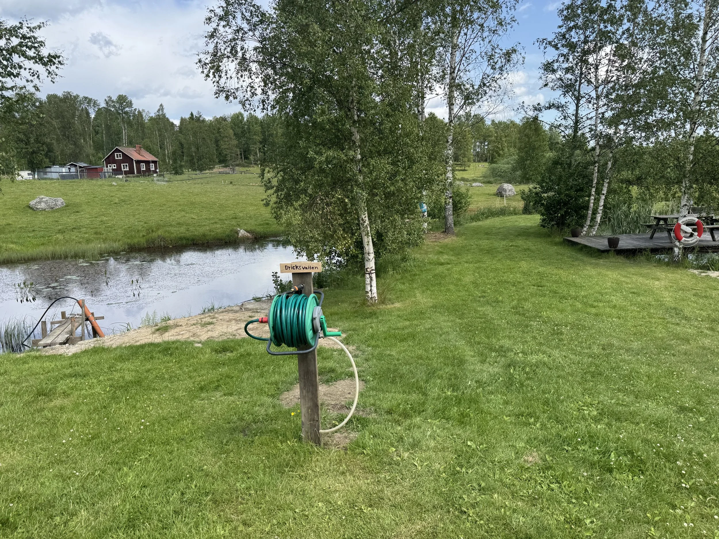 En gräsmatta vid en liten sjö med träd runt omkring. En trädgårdsslang är placerad på marken och det finns en skylt som säger 'Dricksvatten'.