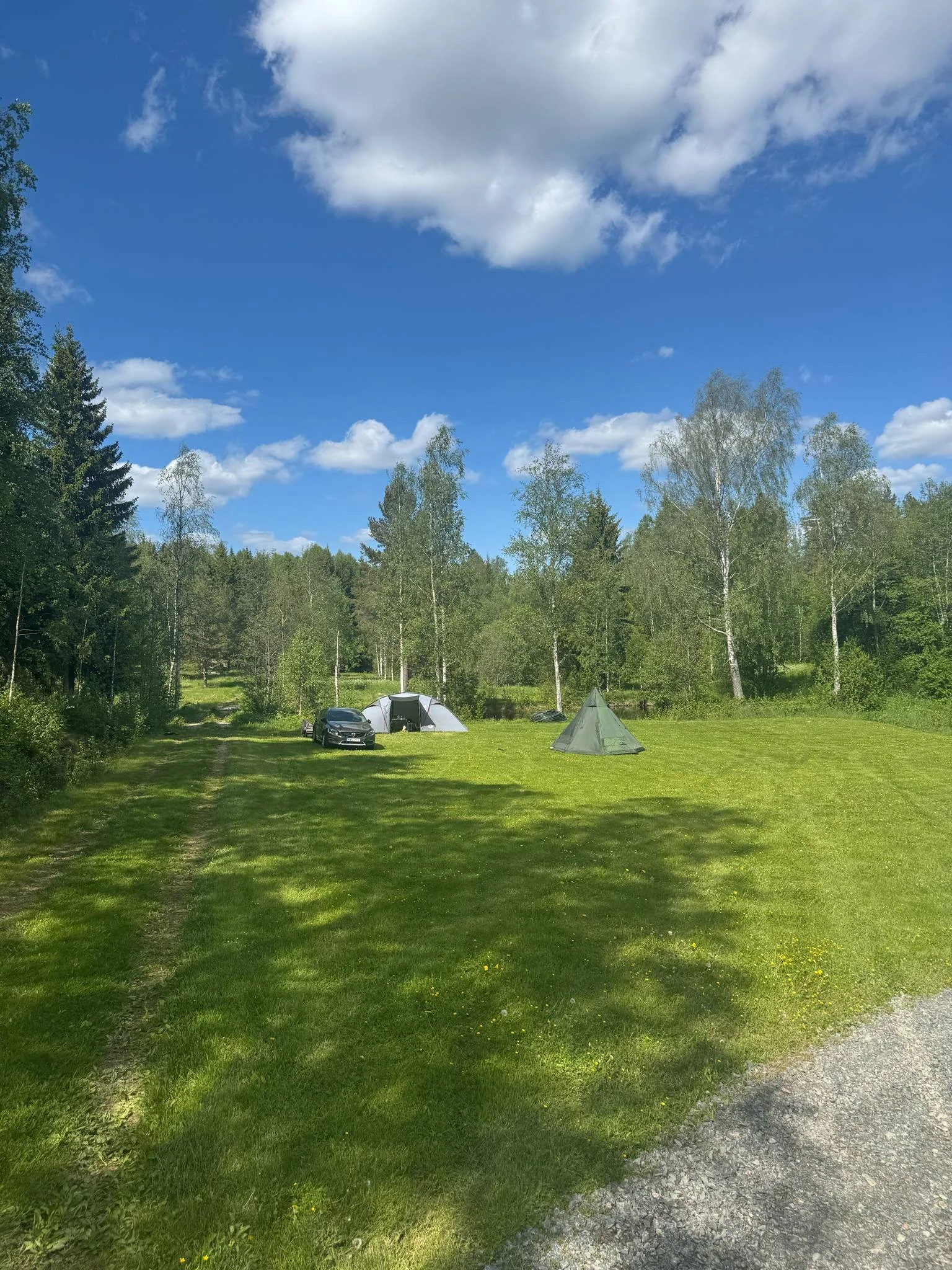 En gräsmatta med två tält, en bil och träd i bakgrunden under en blå himmel med några moln.