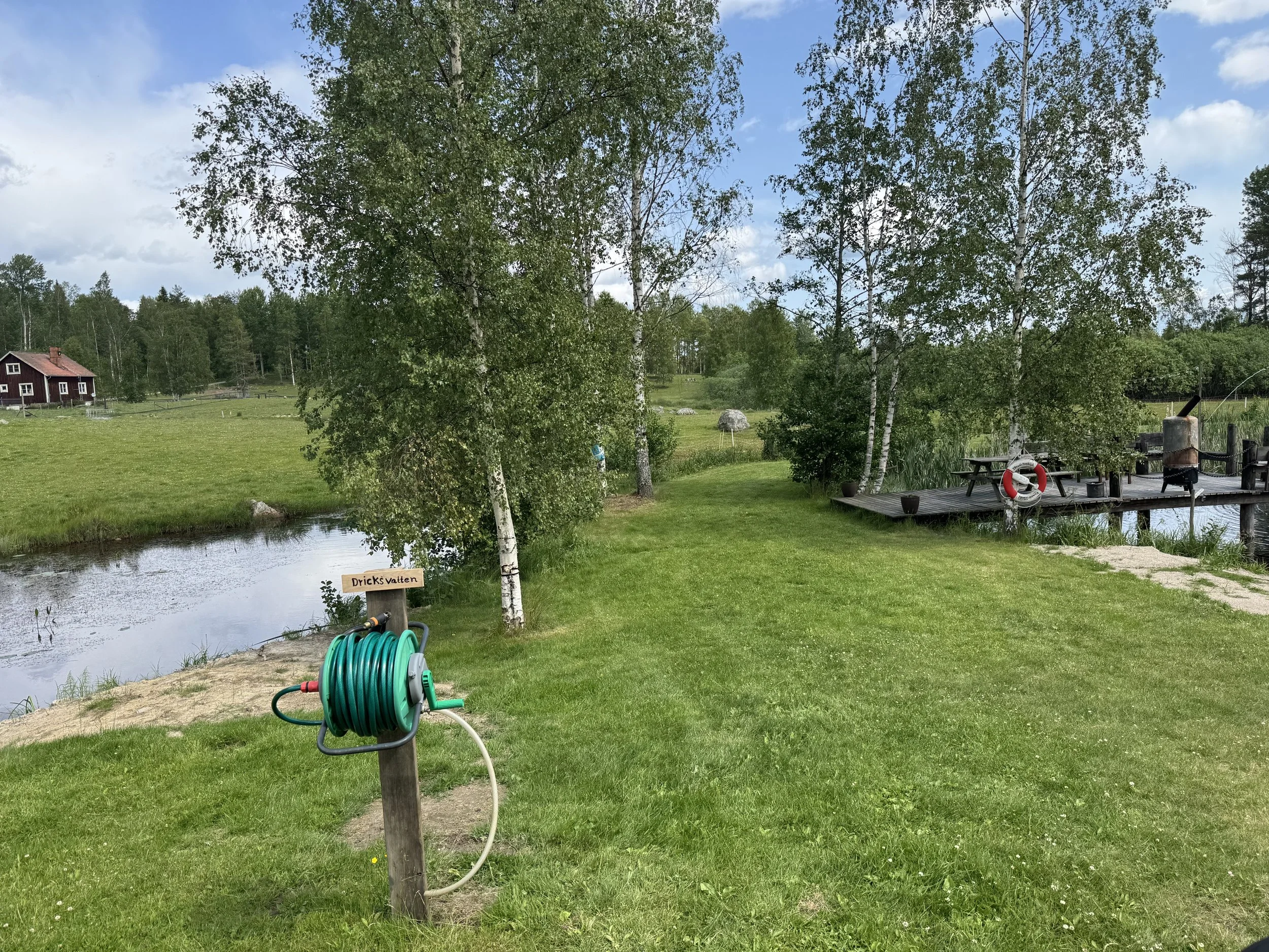 Grön gräsmatta med träd, en liten brygga, vatten och en vindskyddad sittplats i naturen.