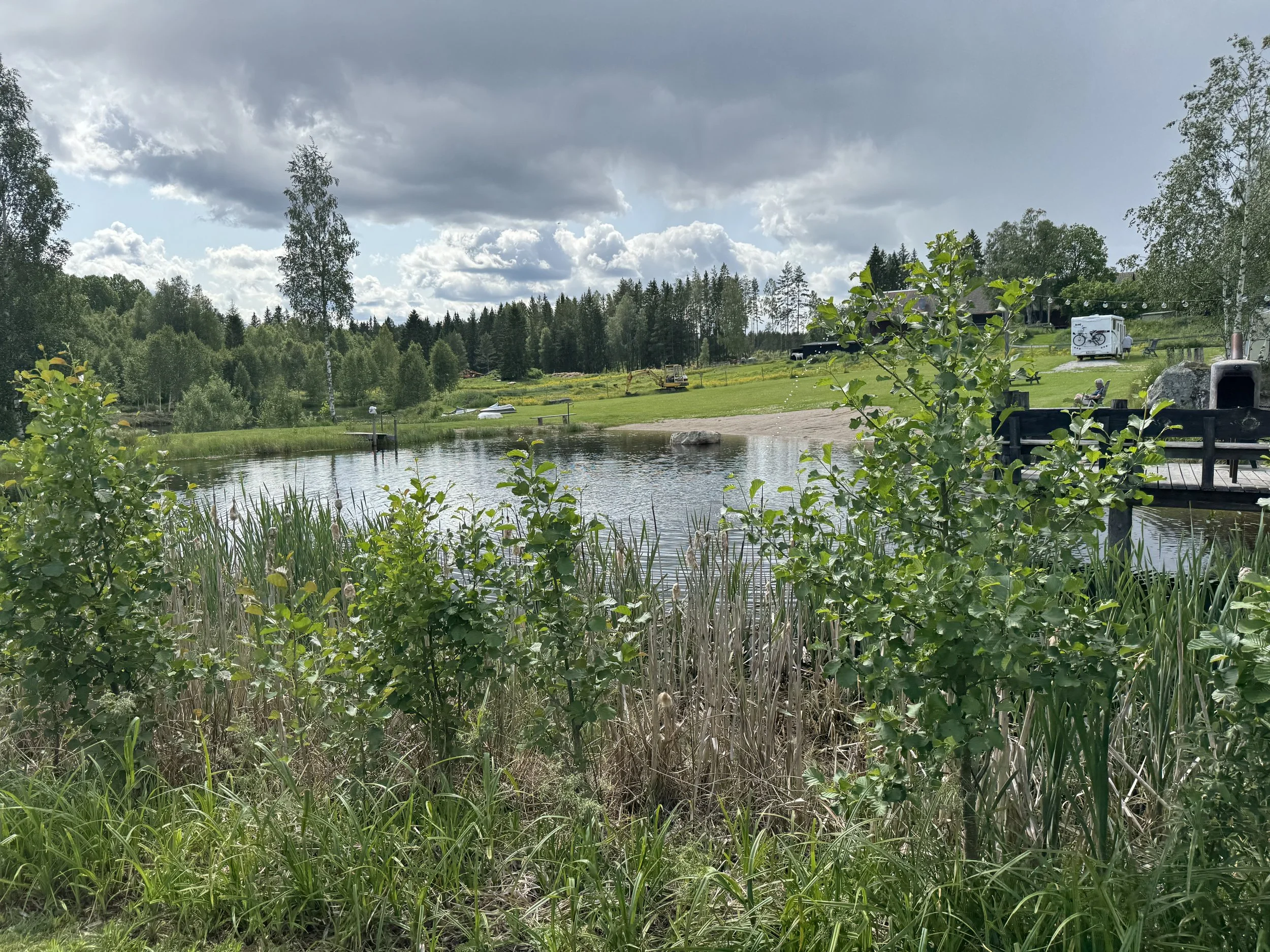 Landskap med en liten sjö, gröna buskar och träd, en bänk vid vattnet och en husvagn i bakgrunden under en molnig himmel.