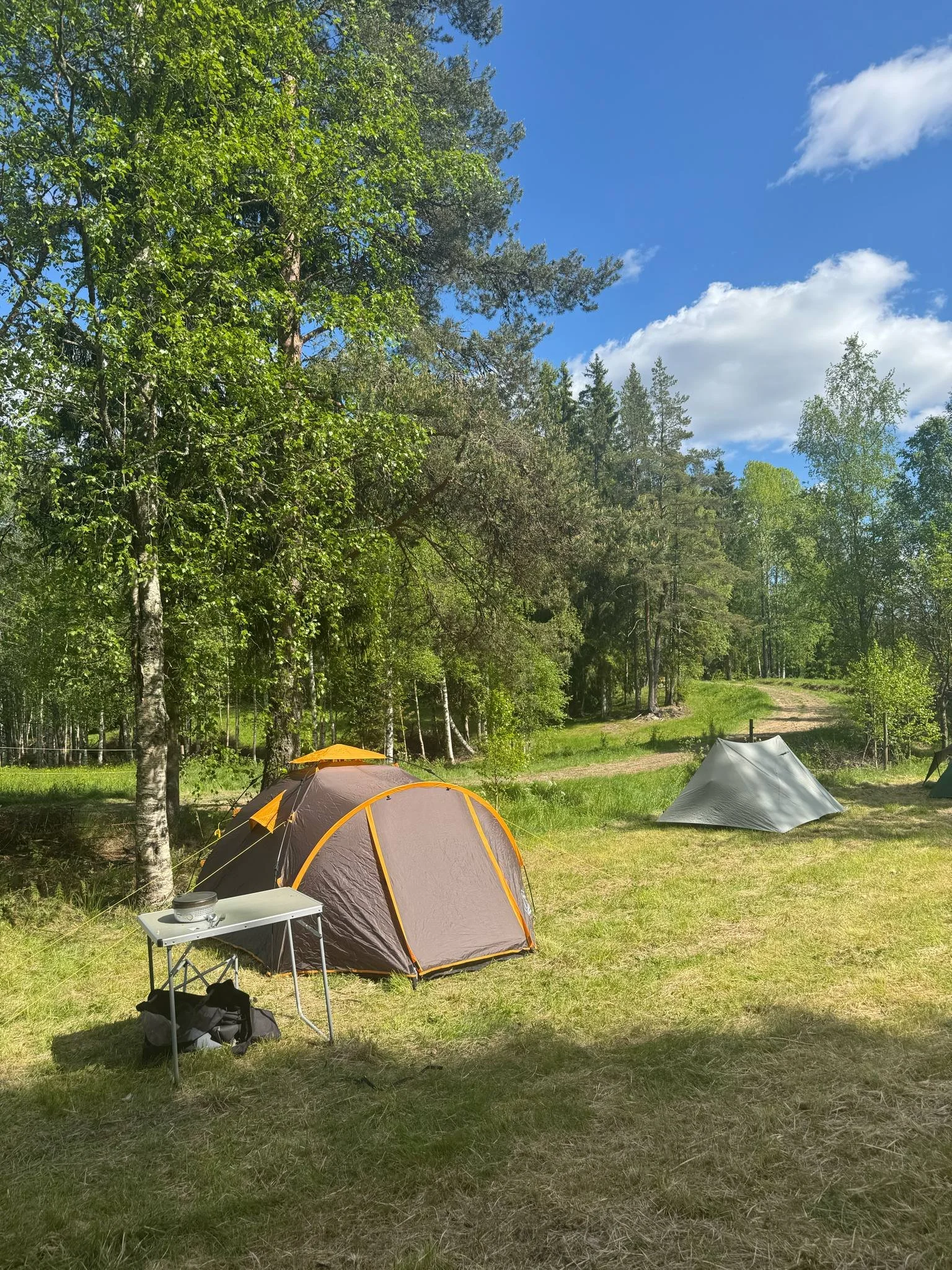 En campingplats med två tält, ett grått och ett grönt, under en klarblå himmel med några moln. Det finns också ett litet bord med saker på, omgivning av gröna träd och gräs.