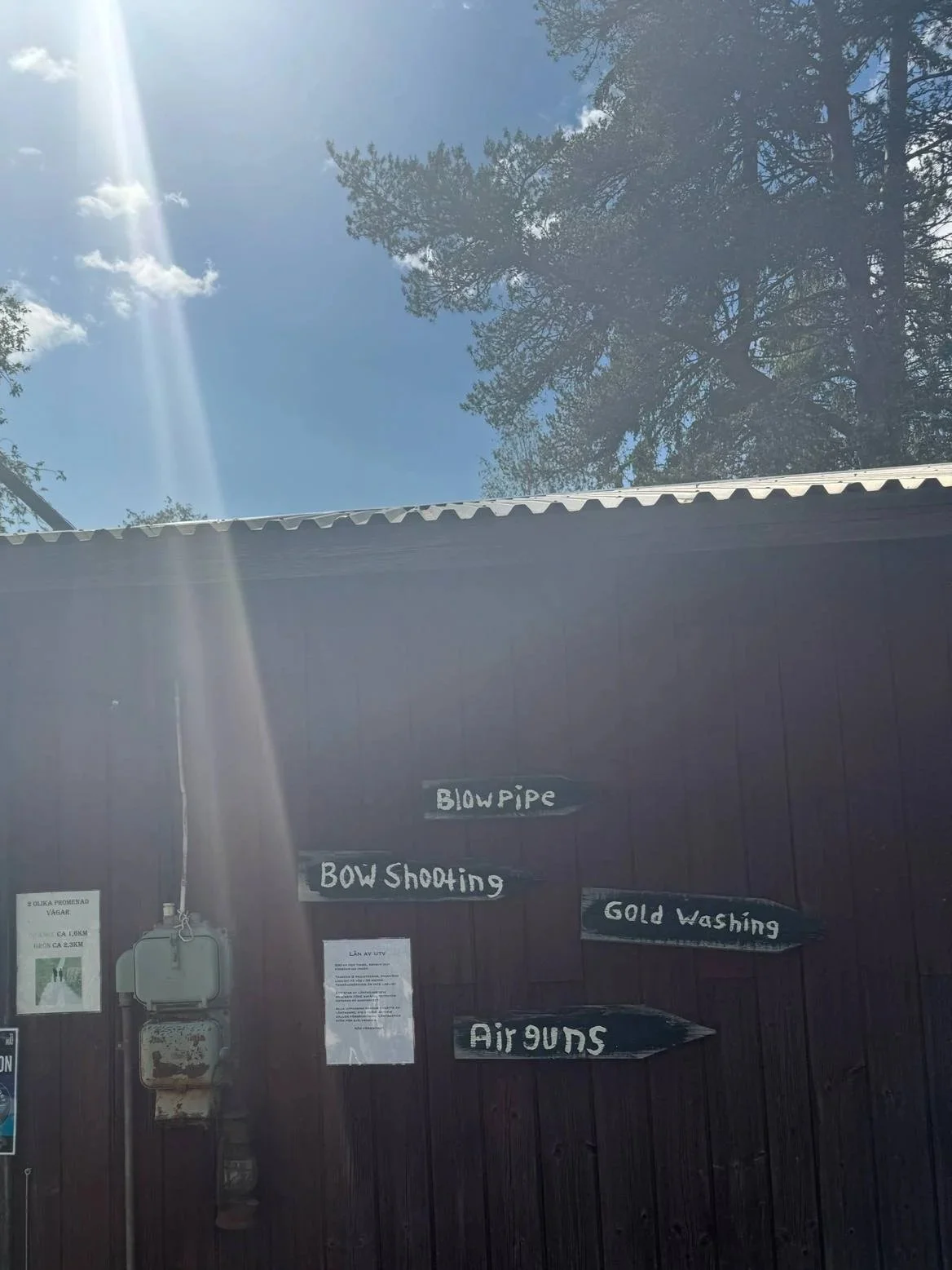 Vägg med skyltar som visar olika aktiviteter: Blowpipe, Bow Shooting, Gold Washing och Air Guns. Solsken och träd i bakgrunden.