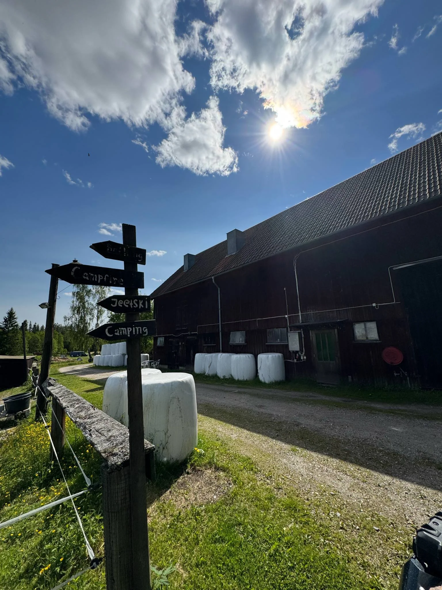 En landskapsbild av en lantlig plats med ett trähus, höbalar utomhus, en grusplan, en träskylt med väderpilar som pekar mot aktiviteter som kan inkludera guidade turer, kajakpaddling, camping och fiske, under en solig himmel med några moln.