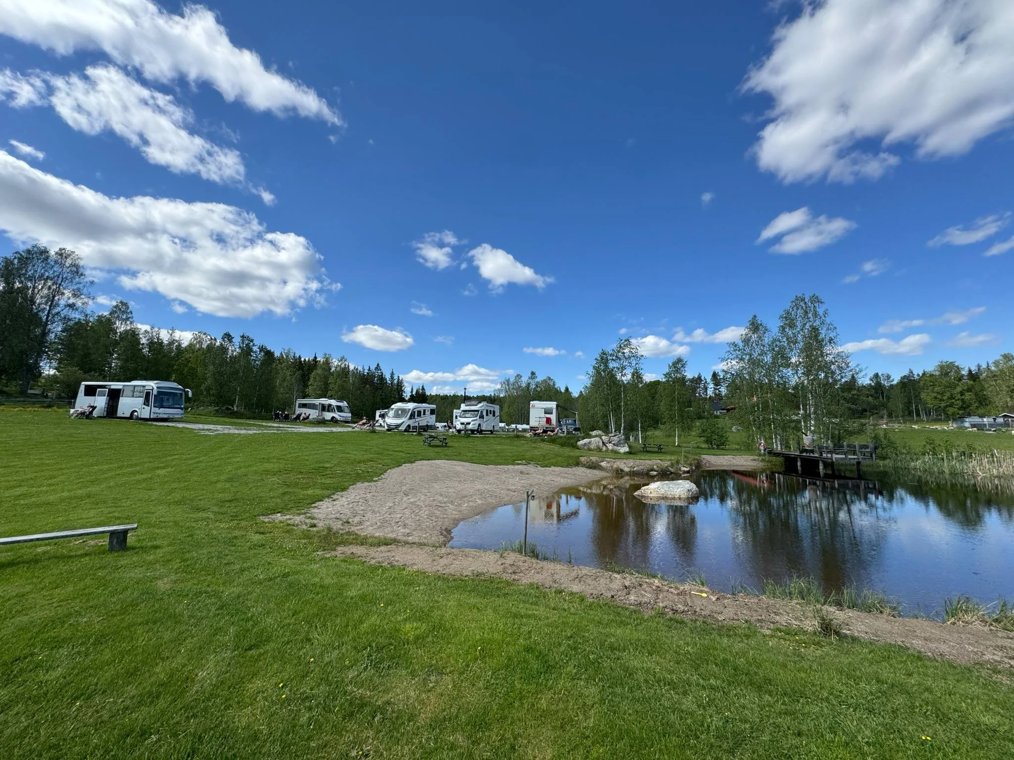 Campingplatser med husvagnar vid en liten sjö under en blå himmel med moln.