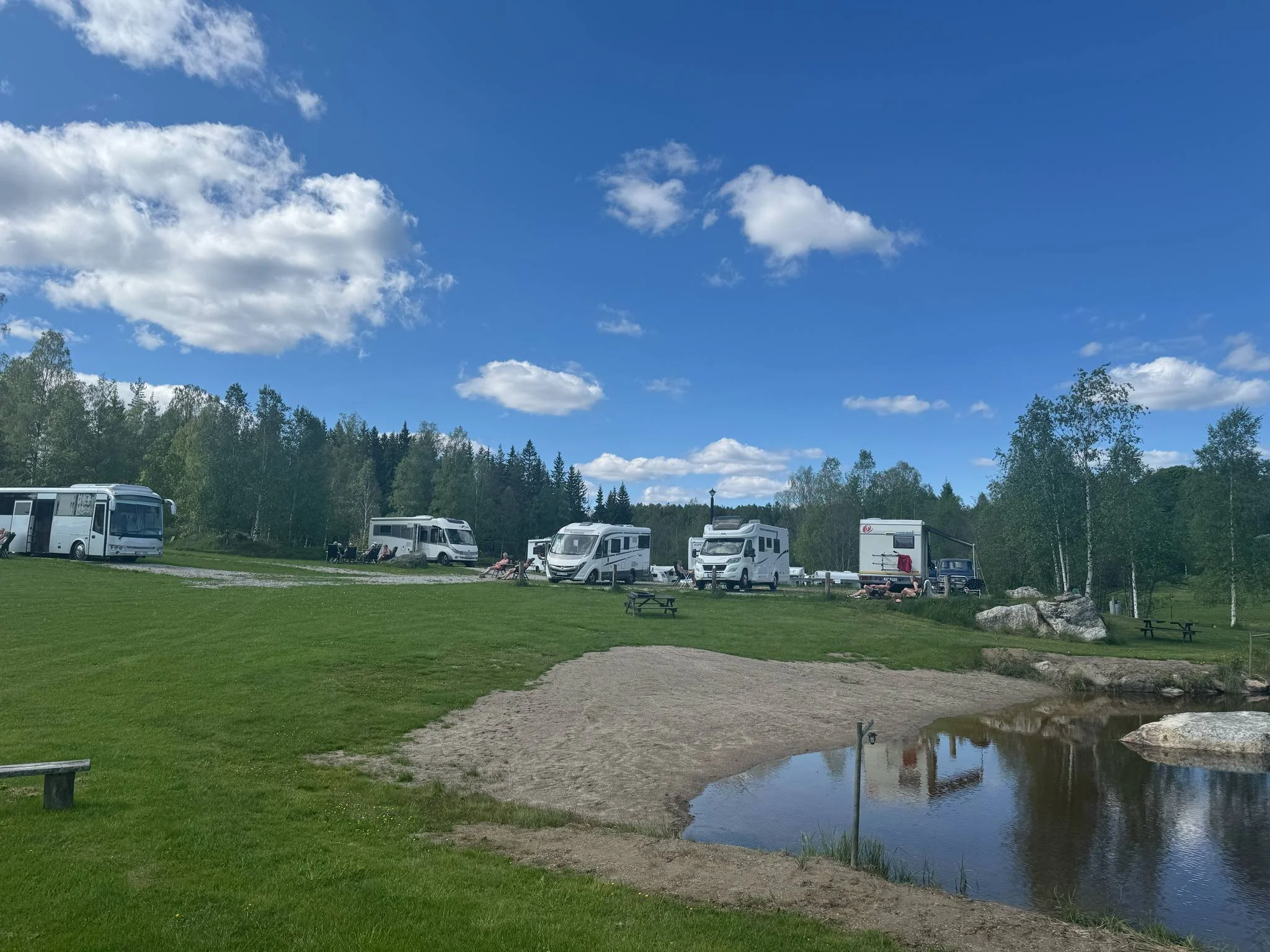 En campingplats med flera husvagnar och husbilar som står parkerade på gräset. Det finns ett litet vatten, en sandstrand och några uteplatser med bänkar. Runt campingen finns träd och en blå himmel med några moln.