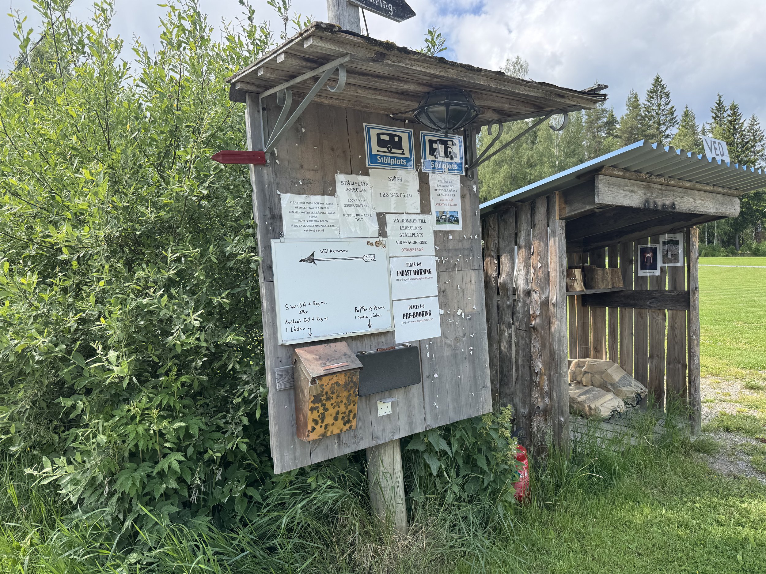 En anslagstavla och ett litet skjul med ved, ute i grönt landskap.
