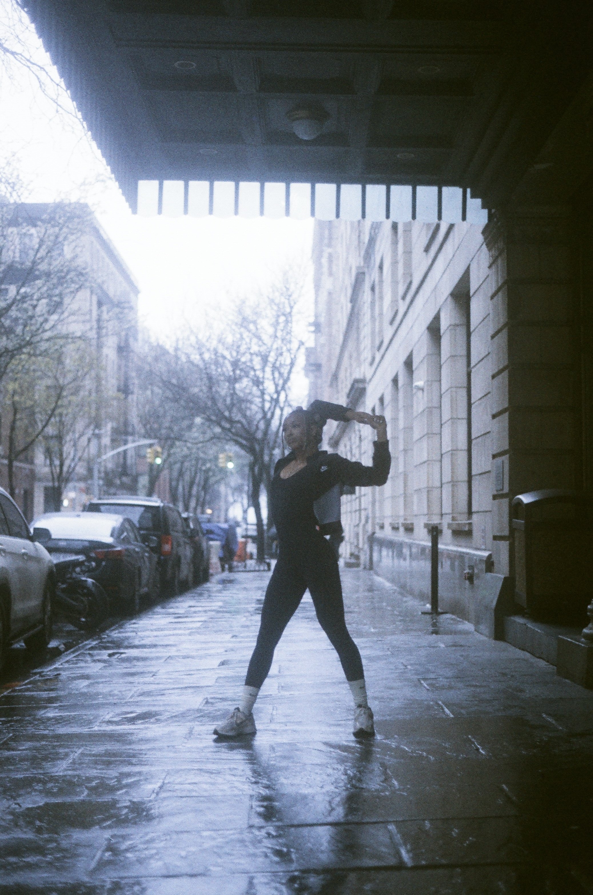 A woman in athletic clothing, standing on a wet sidewalk outdoors, holding her hair up with one hand, with a row of parked cars and leafless trees in the background.