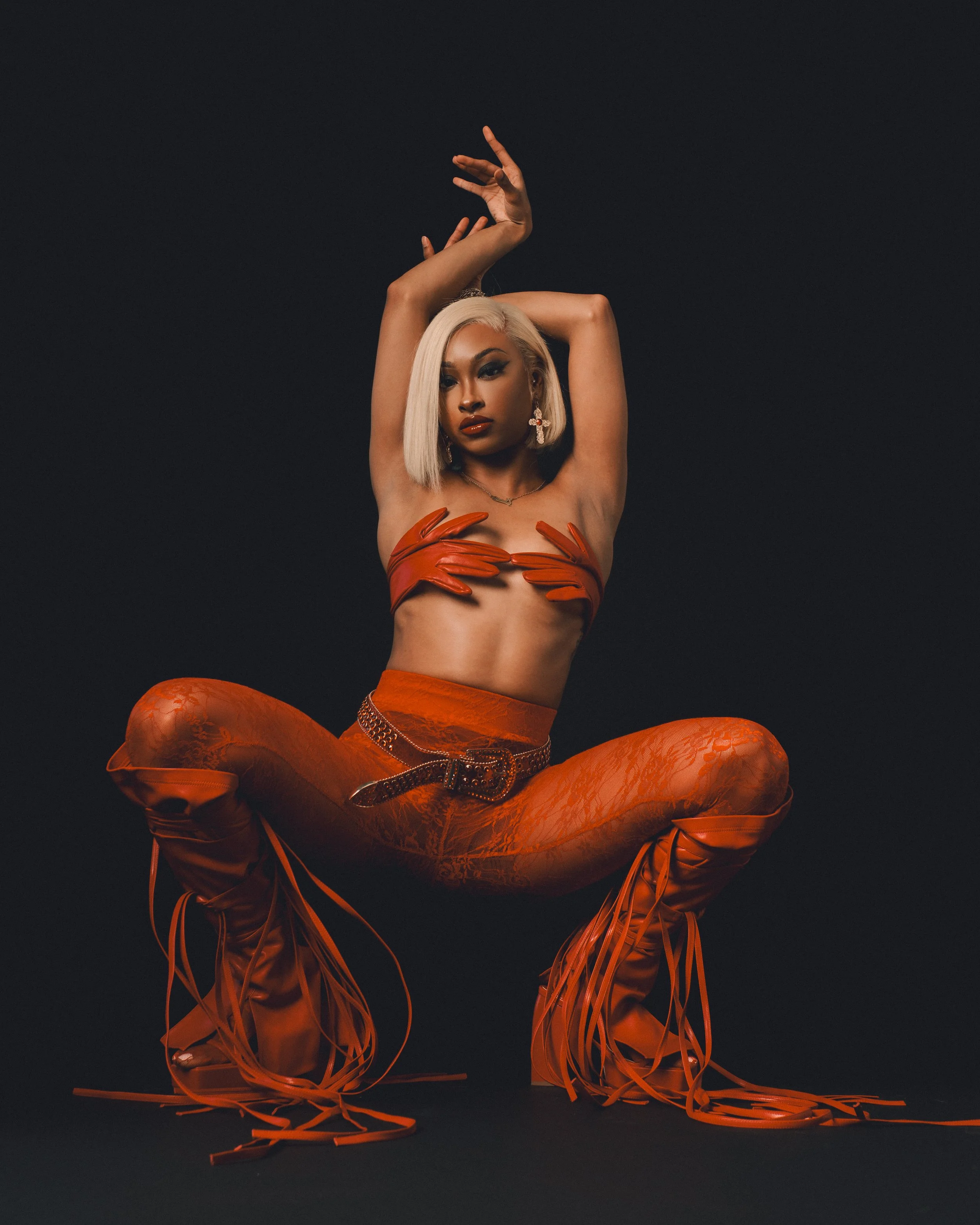 A woman with platinum blonde hair, dressed in orange lace leggings, thigh-high lace-up boots, and orange gloves, strikes a pose with arms raised above her head against a dark background.