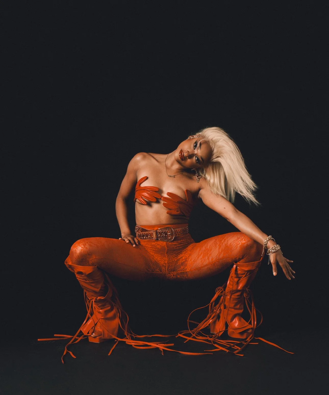Woman with platinum blonde hair, wearing orange leather pants and high laced boots, posing against a black background.