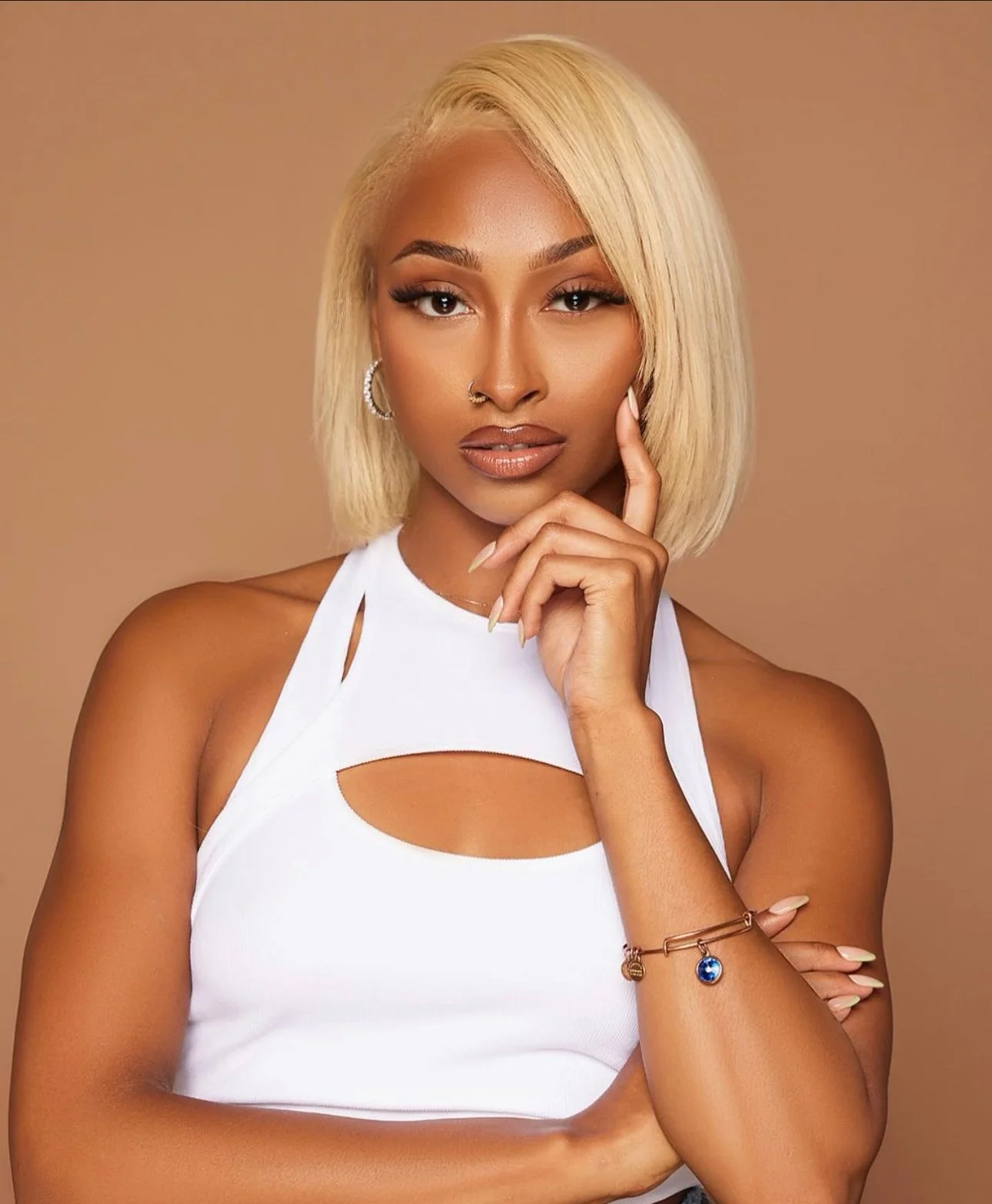 A woman with blonde hair, wearing a white top with cutouts, posing with her hand near her face against a neutral background.