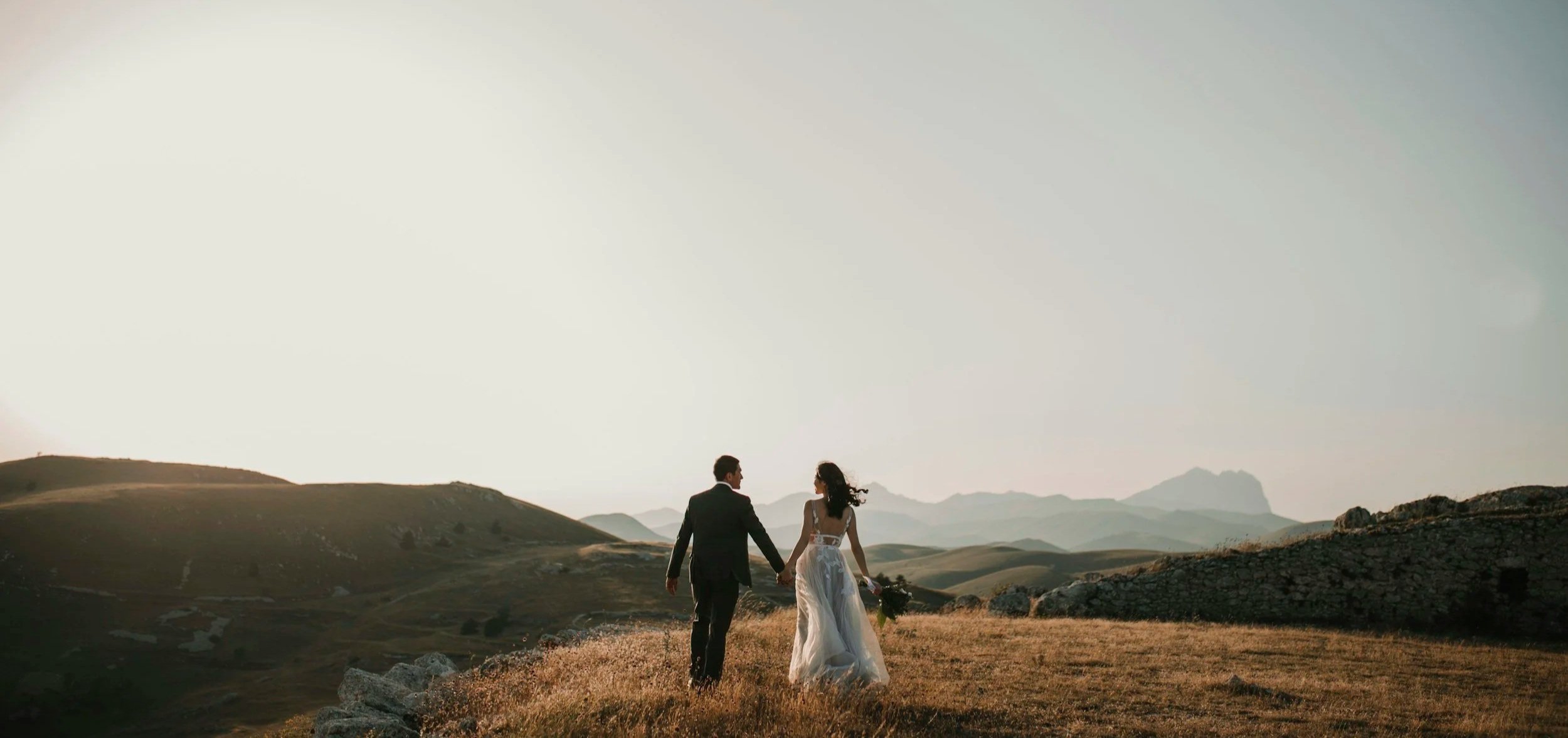 A couple in wedding attire walking hand in hand across a grassy, hilly landscape at sunset or sunrise.