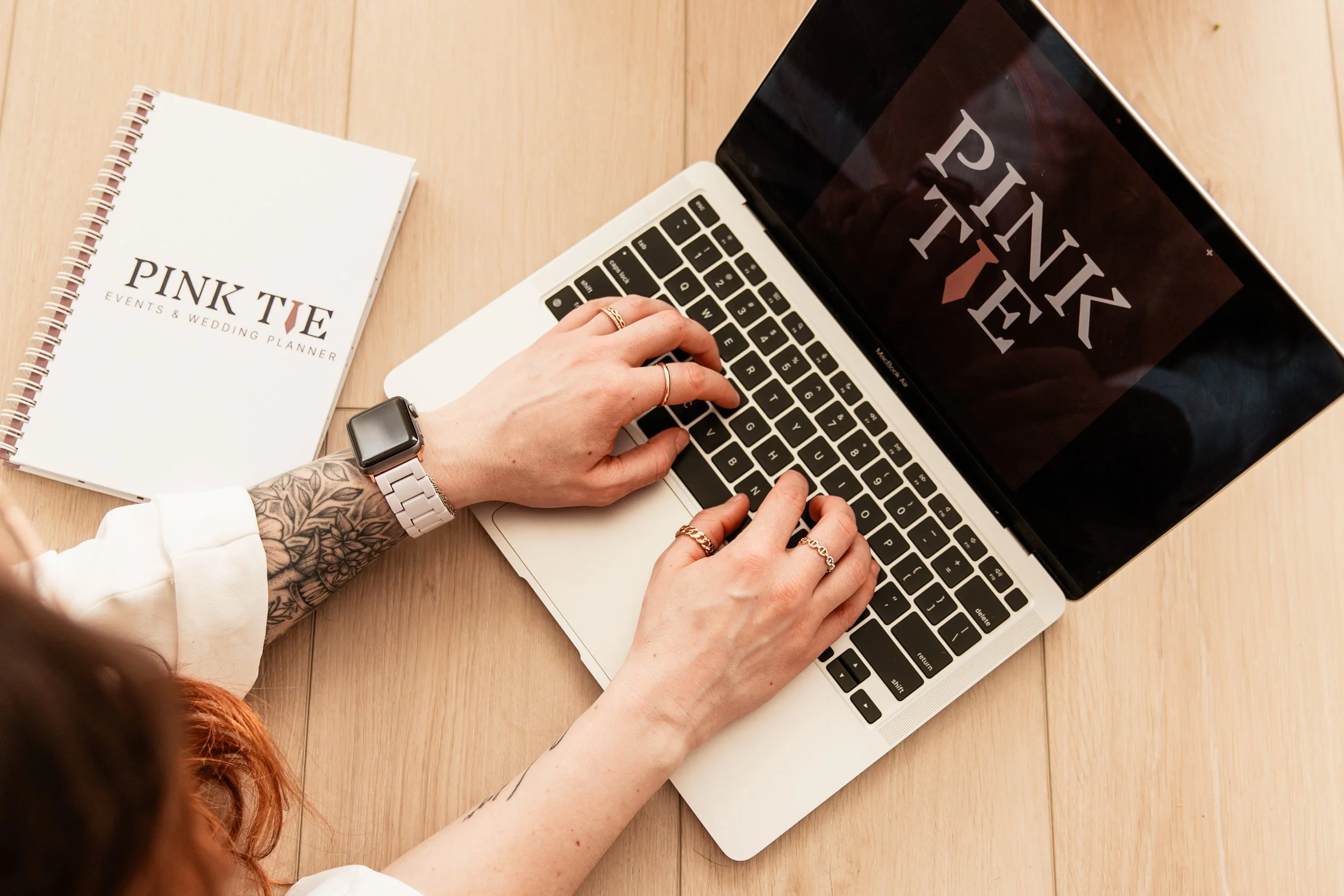 A person with tattoos on their arm, wearing a smartwatch and rings, typing on a silver MacBook laptop. Next to the laptop is a spiral-bound planner with the title 'PINK TIE EVENTS & WEDDING PLANNER' on a wooden surface.